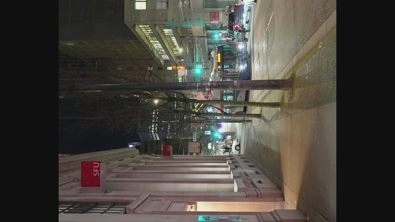 The image depicts a city street scene at night, likely in Vancouver, Canada, given the presence of a "SFU" (Simon Fraser University) sign. The perspective is from a sidewalk, looking down a street lined with trees and buildings.
On the left, a cream-colored building with classical architectural details features a prominent red sign with "SFU" in white lettering. Further down the sidewalk, another smaller "SFU" sign is visible. A vertical sign with the number "640" is illuminated near the left edge of the frame. The sidewalk itself is paved and shows some scattered debris.
The street is active with vehicle traffic. A blue and white trolley bus, labeled with "14 HASTINGS" and a route number, is in the center of the road. To its right, a dark-colored pickup truck is driving, and further ahead, a white sedan is parked or moving. Other vehicles are visible in the distance.
Pedestrians are present on the sidewalk, though their details are indistinct due to the low light conditions and distance. Traffic lights are visible in the background, indicating an intersection. The environment suggests a commercial or institutional area within the city, illuminated by streetlights and building lights. The wet appearance of the street indicates it may have recently rained.
