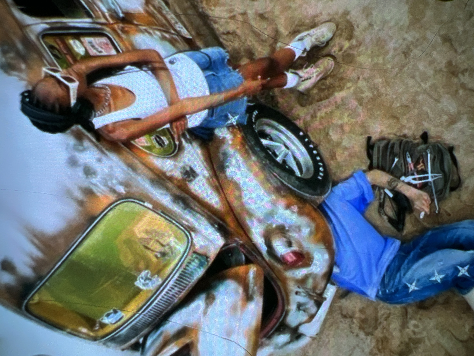 This is a high-angle shot of a young Black woman and a young Black person, possibly male, interacting in an outdoor urban environment that is implied to be Lagos, Nigeria. The woman is standing and appears to be posing next to a vintage Volkswagen Beetle that has been customized with an airbrushed, distressed, and colorful design. She is wearing a white tank top, a denim skirt, white sunglasses, and white sneakers. The other person is lying on the ground in front of the car, wearing a blue t-shirt and blue jeans with white star patterns. They are positioned near a backpack. The ground appears to be sandy or dirt-like, consistent with a dry, outdoor setting that could be found in Lagos. The lighting suggests it is daytime.