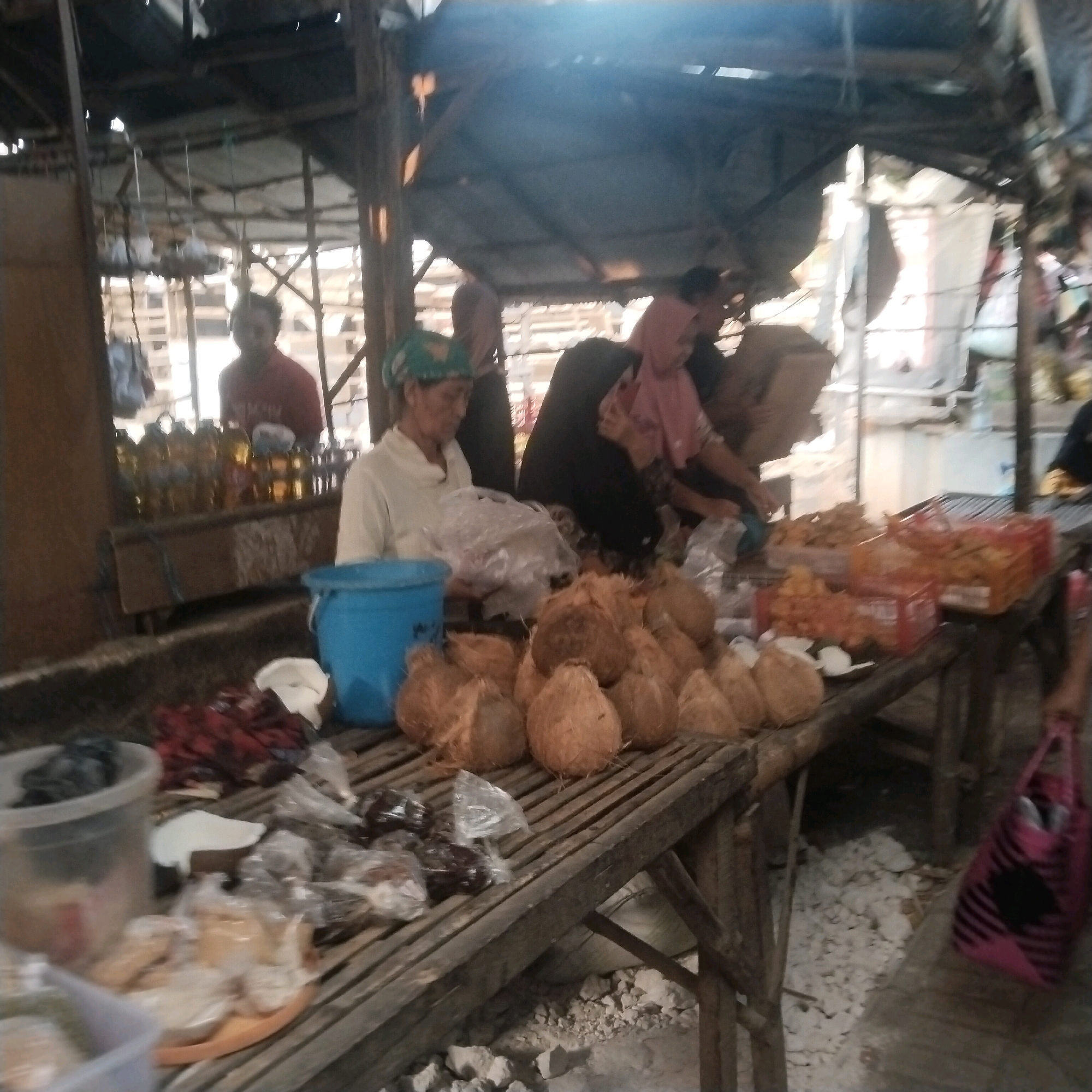 The image depicts a bustling marketplace scene, likely in Kesek, Indonesia. In the foreground, a wooden stall is laden with goods. A pile of coconuts is prominent, some whole and some halved. Various food items are displayed in plastic bags and containers, some appearing to be fried snacks. To the left, a clear plastic tub holds more food items. A blue bucket is also visible on the stall.
In the mid-ground, several individuals are present. One woman, wearing a green headscarf and a light-colored top, is attending to the stall. She appears to be interacting with a customer or arranging goods. Another woman, wearing a pink headscarf and a patterned dress, is also visible behind the stall. Further back, a person in a red shirt is standing. The marketplace has a rustic, open-air structure with a corrugated metal roof, suggesting a covered but not fully enclosed space. The overall impression is one of local commerce and community activity.