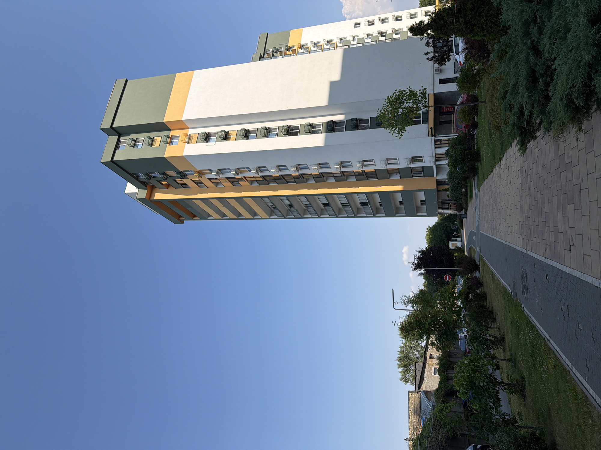 The image depicts a modern, multi-story apartment building with a distinctive facade featuring bands of white, yellow, and dark green. The building is situated in a residential area with a clear blue sky overhead. In the foreground, a paved walkway runs alongside a green lawn with various shrubs and trees. Several parked cars are visible near the building, and a street lamp stands along the pathway. In the background, another similar building is partially visible, and further back, a low-rise brick structure and trees suggest a developed urban environment. The scene appears to be a quiet, well-maintained residential complex.