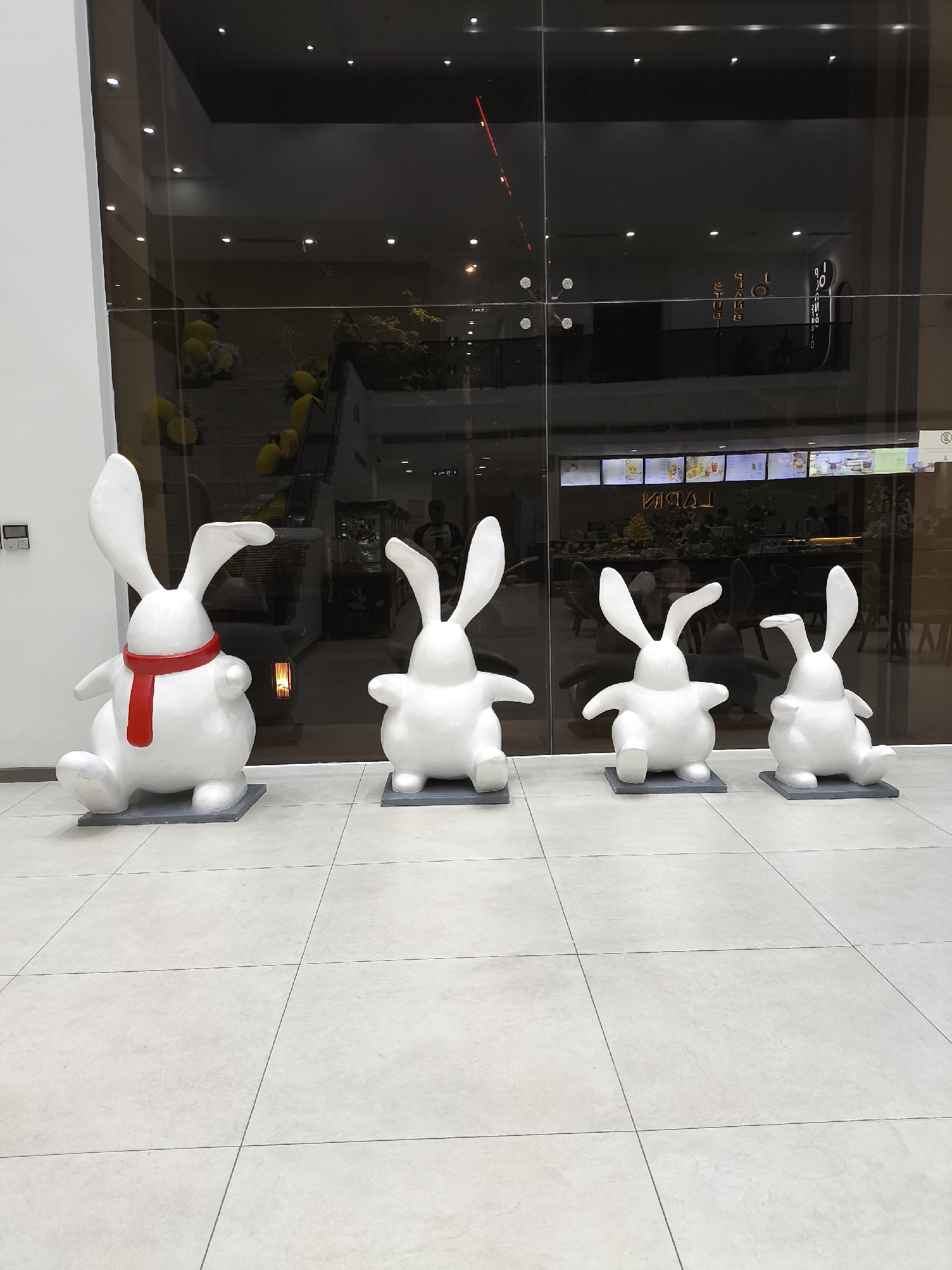 This media file captures a display of four white, cartoonish rabbit statues arranged in a line on a tiled floor. The largest rabbit, on the left, is adorned with a bright red scarf. The statues are positioned in front of a large glass-paneled wall that reflects the interior of what appears to be a shopping mall or a commercial establishment.
Through the glass, the reflection reveals a dimly lit space with rows of illuminated display boards, likely showcasing menus or products. Yellow balloons are visible in the background, suggesting a festive or decorative theme. The overall atmosphere is calm and still, with no visible activity or people interacting with the statues. The lighting suggests it could be daytime, but the interior setting makes it difficult to ascertain the exact time of day.
The scene is set indoors, on a light-colored tiled floor with a grid pattern. The building appears modern, with a sleek glass facade. There are no discernible weather cues, and the emotion conveyed is one of whimsical display rather than active engagement. The text visible through the glass appears to be in a foreign language, possibly Vietnamese given the location context of Đà Nẵng, Vietnam. The words "TABAL" are visible, mirrored due to the reflection.