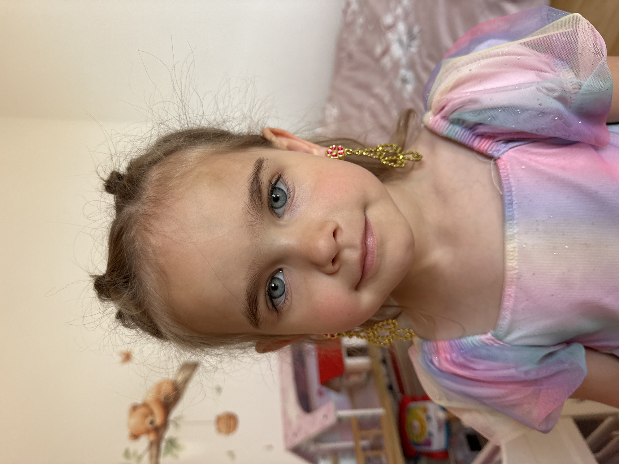 A young girl is looking at the camera and smiling. She has fair skin and light hair pulled into small buns. She is wearing gold earrings and a multi-colored pastel dress. A pink bed and a wall with a squirrel and leaves sticker are visible in the background. The location is likely a home, but not Niederquembach, Germany.