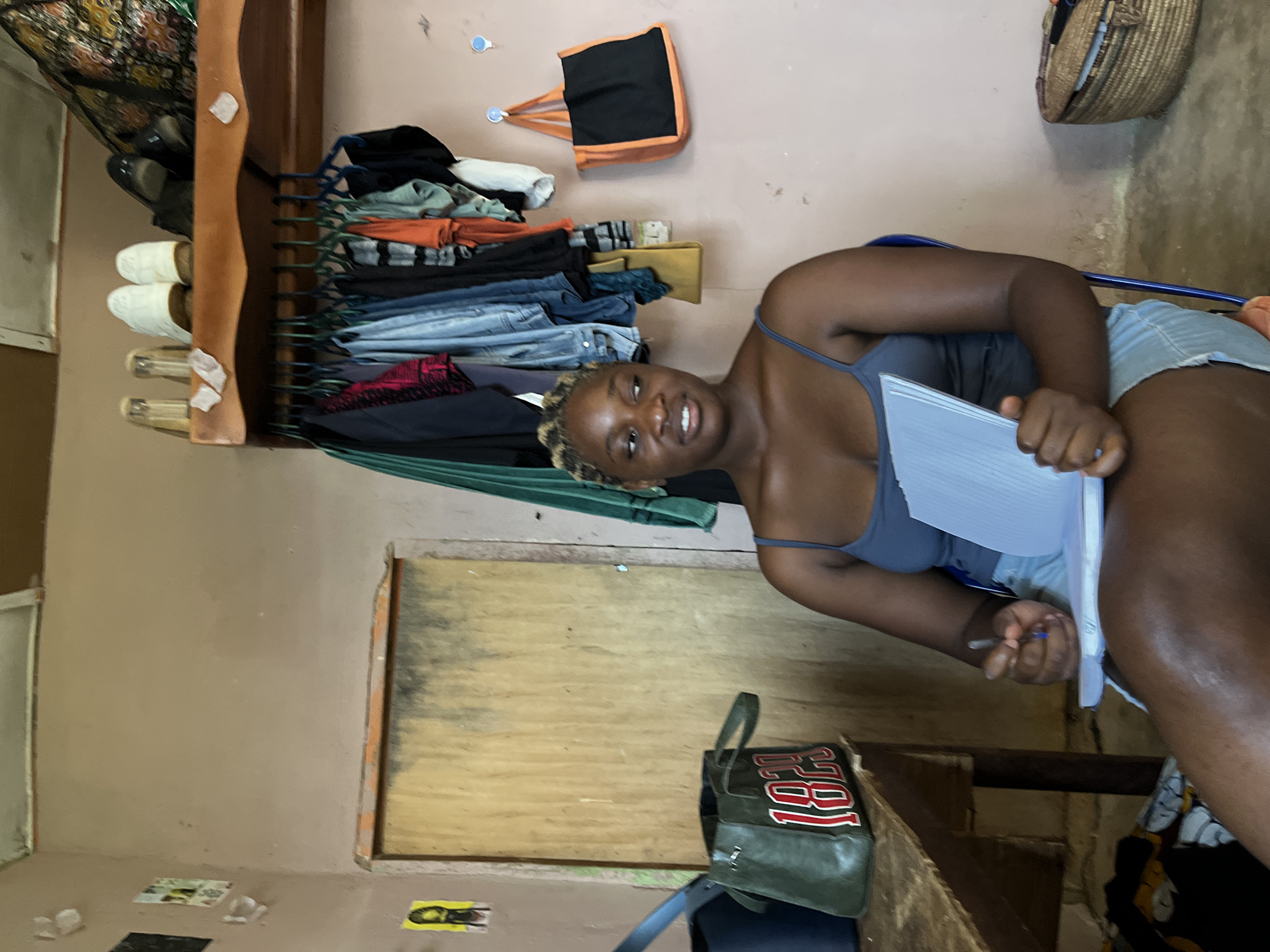 In Rafin Par, Nigeria, a young Black woman is seated in a blue chair, holding a notebook and pen, seemingly engaged in writing or studying. She wears a gray tank top and denim shorts. The room appears to be a personal living space, possibly a bedroom, with a light pink wall as the backdrop. To the left of the woman, a wooden table is visible, adorned with a green bag displaying the number "1829" in red and white. Above the table, a shelf holds several pairs of shoes and glasses. A wooden door with vertical planks is positioned behind the woman. The wall to her right is decorated with hanging clothes on hangers and a black and orange tote bag. A woven basket sits on the floor to the far right of the frame. The overall impression is one of a simple, functional room.