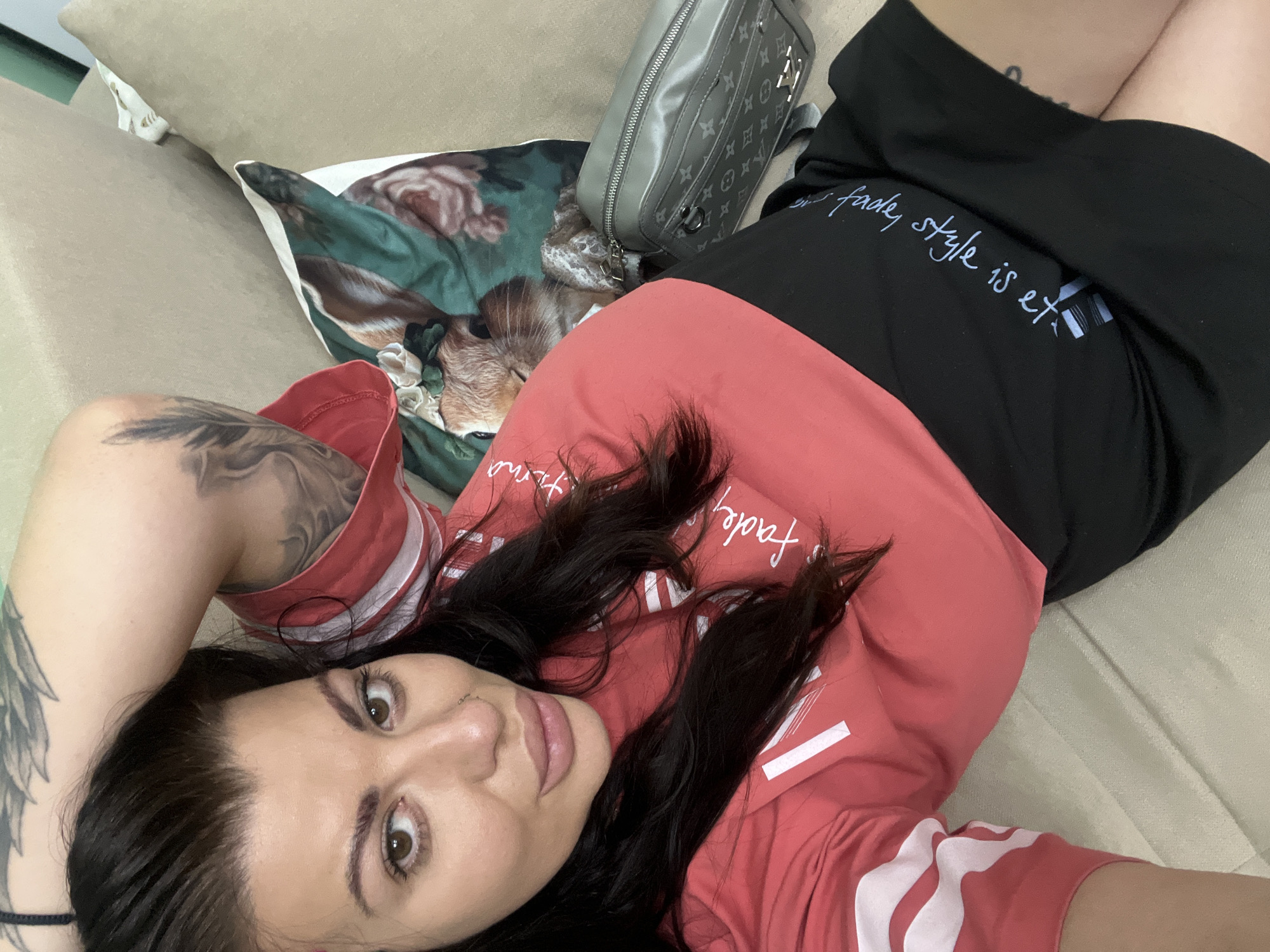 A young woman is lying on a sofa in a domestic indoor setting. She has dark hair and is wearing a red and black outfit. Her arm is raised behind her head. To her right, there is a patterned cushion and a grey bag. The background consists of a beige sofa and wall. The image appears to have been taken in a residential area of Plovdiv, Bulgaria.