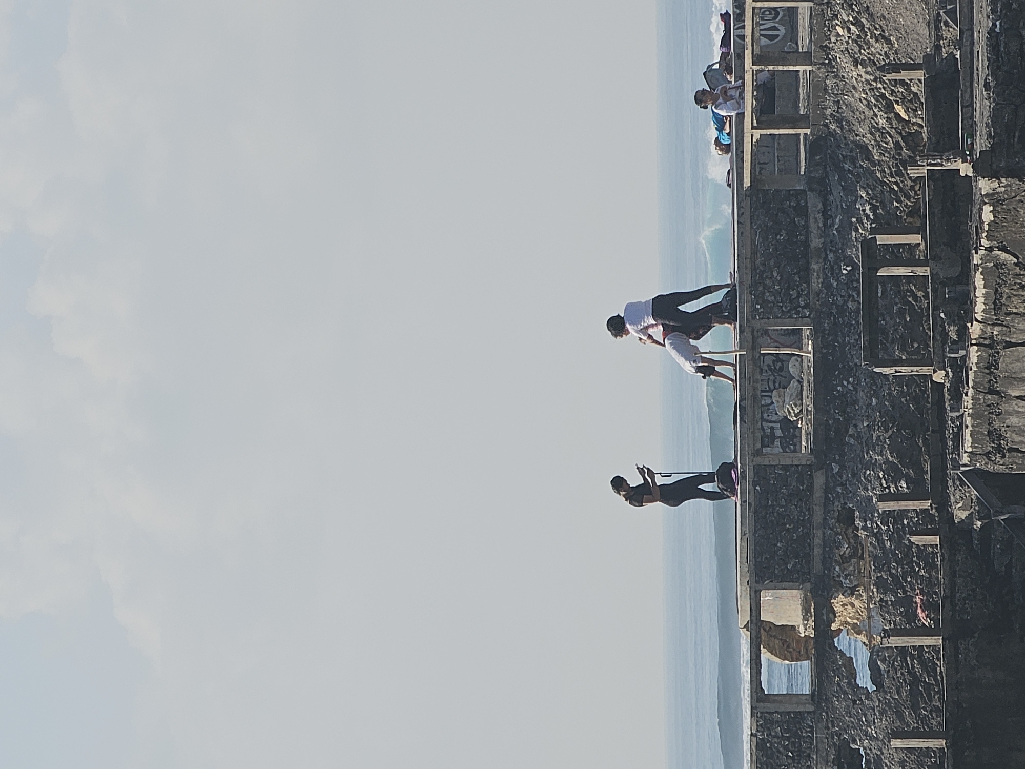 The image shows a group of people on a dilapidated concrete structure overlooking the ocean. The sky is overcast and the water is a pale blue.
On the structure, there are several individuals. Two women are in the process of a yoga pose, with one assisting the other. To the left, a solitary woman is standing and looking at her phone. To the right, two people are lying down on mats, one of whom is wearing a blue shirt and the other a white shirt. There are also some bags and other items scattered on the structure.
The structure itself appears to be abandoned and in disrepair, with exposed rebar and crumbling concrete. There are some graffiti tags visible on the wall. The ocean behind the structure has visible waves rolling in.
This scene appears to be located in Denpasar, Indonesia, likely on the coast. The overall impression is one of an outdoor recreational activity taking place in a somewhat neglected but scenic environment.