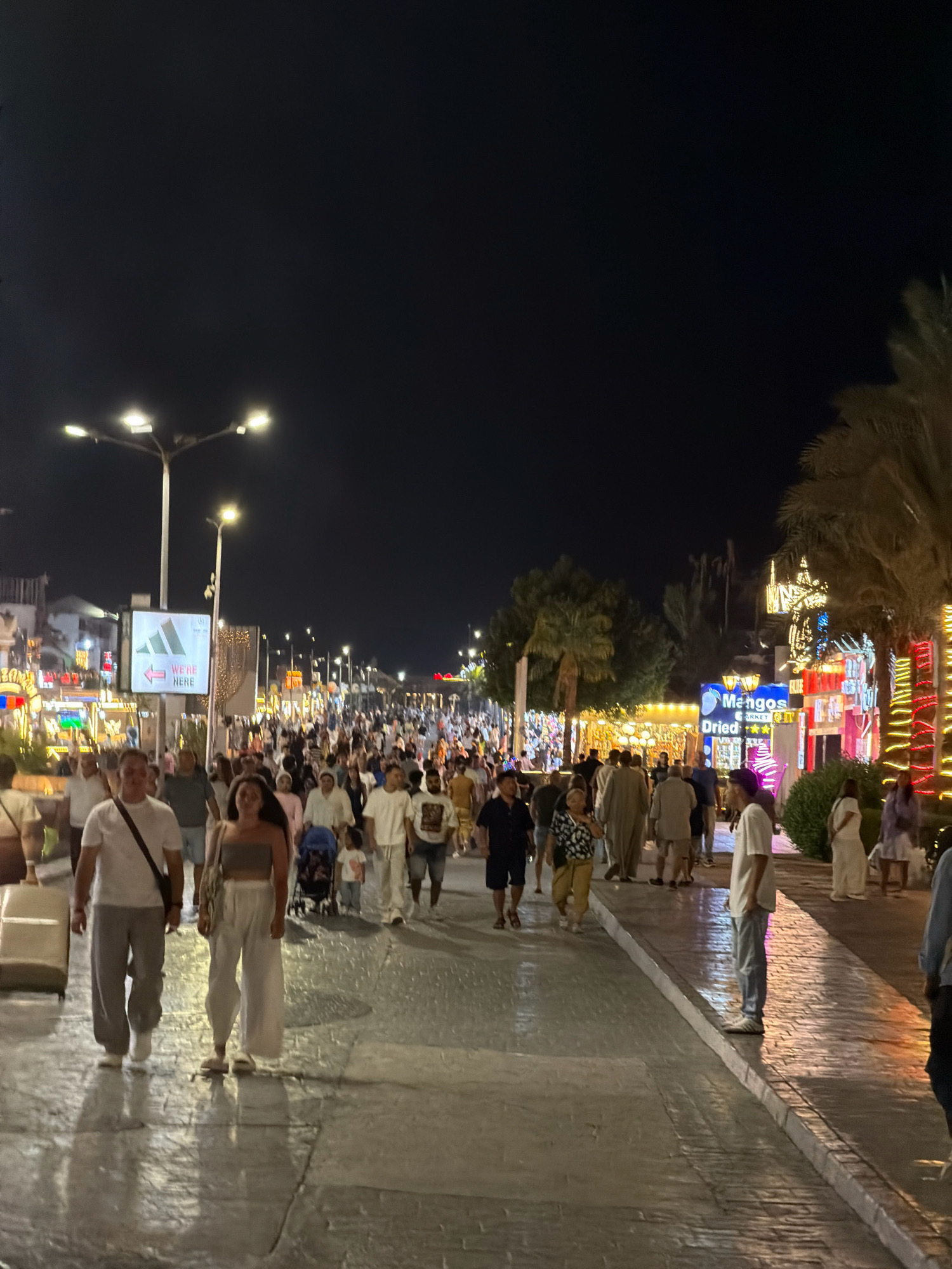 The image depicts a bustling street scene at night in Sharm El Sheikh, Egypt, likely on Naama Bay promenade, a popular tourist area. A diverse crowd of people are walking along a wide, paved walkway lined with shops and restaurants adorned with bright lights and festive decorations, including palm trees wrapped in colorful lights. Several individuals are visible in the foreground, including a man and a woman walking side-by-side, a man with a suitcase, and other pedestrians moving through the crowd. The street is illuminated by streetlights and the vibrant glow from the businesses, creating a lively and energetic atmosphere. The dark night sky provides a backdrop for the illuminated scene.