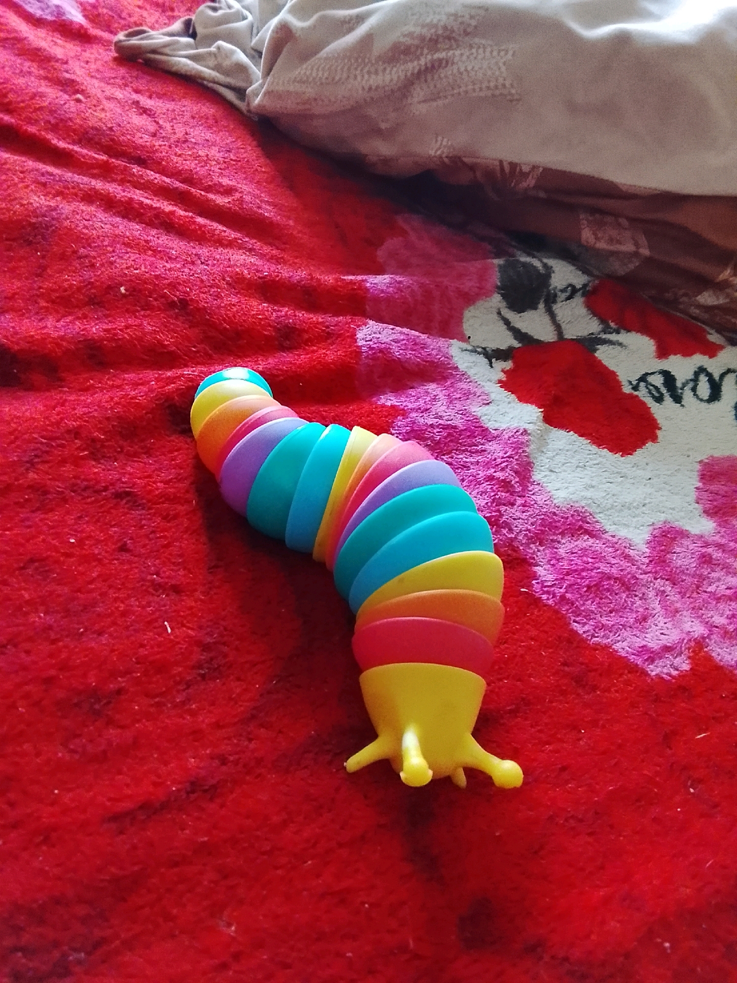 A brightly colored, segmented toy caterpillar lies on a textured red surface, possibly a blanket or carpet. The toy is composed of multiple curved segments in various shades of yellow, orange, pink, purple, and blue, forming a flexible, snake-like shape. The front of the toy tapers to a yellow head with two small, protruding legs or antennae. In the background, a folded piece of fabric, lighter in color and with a textured pattern, is visible, suggesting a domestic setting. The red surface beneath the toy has a slightly fuzzy texture and appears to be a solid color with some subtle darker variations, possibly from shadows or the texture itself. The overall lighting suggests an indoor environment. There is no indication of the city of Karma, Belarus.