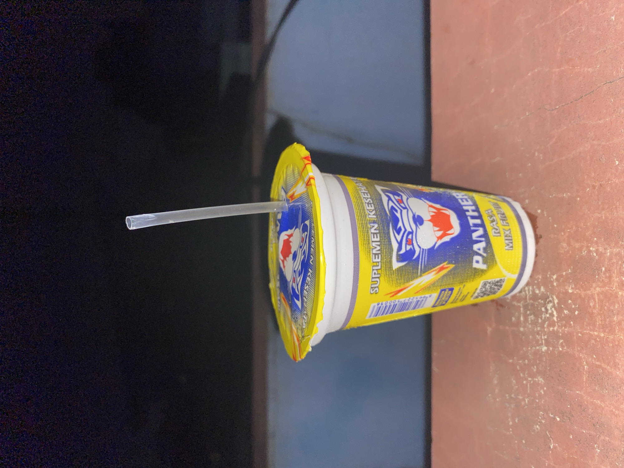 A close-up, eye-level shot captures a single, disposable cup filled with a beverage, its yellow foil lid partially peeled back to reveal a clear plastic straw inserted into the drink. The cup is primarily white with a vibrant yellow label featuring a blue and red panther logo and text in Indonesian, including "SUPLEMEN KESEHATAN" (Health Supplement) and "PANTHER". Below the panther logo, smaller text reads "RASA MIX FRUIT" (Mixed Fruit Flavor). The cup rests on a textured, reddish-brown surface that appears to be an outdoor table or ledge. In the background, a dark, blurry expanse suggests a nighttime setting, possibly outdoors in Tangerang, Indonesia. The lighting is dim, with the cup itself being the primary illuminated subject, highlighting the condensation on its surface.