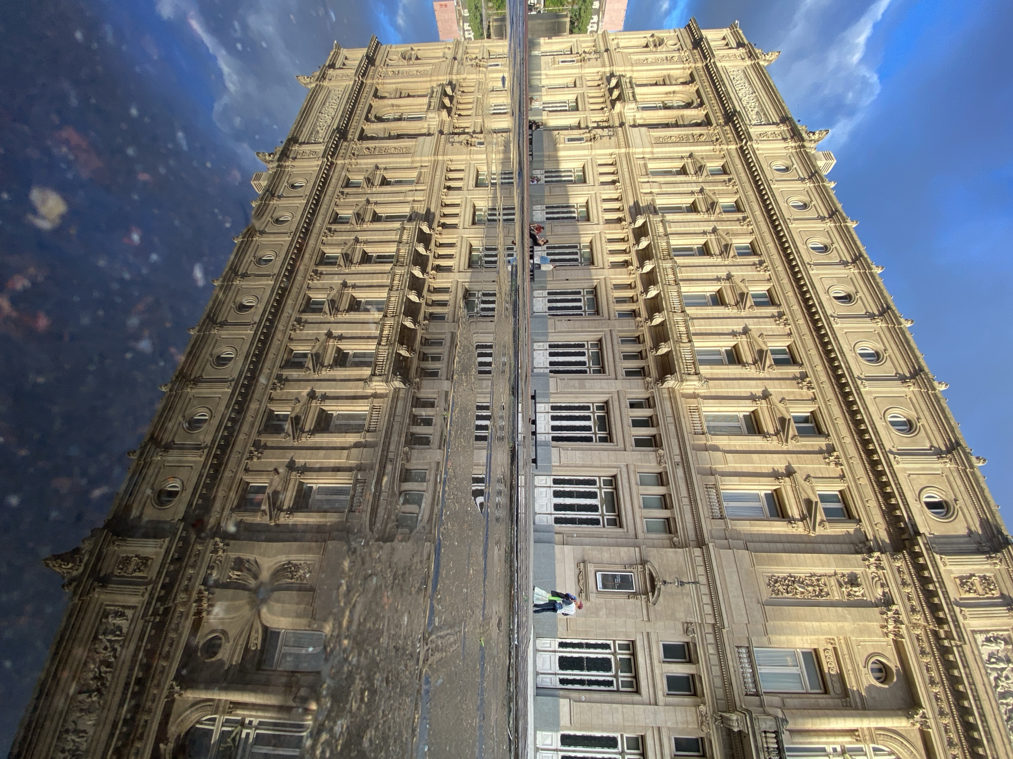 This unique image presents an ornate building in Buenos Aires, Argentina, captured through its reflection in a large puddle on the ground. The photograph is oriented vertically, with a central horizontal line dividing the actual scene (lower half) from its inverted reflection (upper half).
The main subject is a grand, multi-story building featuring classical architecture. Its façade is constructed from light-colored stone, possibly beige or tan, adorned with intricate carvings, decorative motifs, and numerous windows of various shapes, including rectangular panes and circular portholes. The building's imposing structure and detailed craftsmanship suggest a historic and prominent landmark.
In the lower portion of the image, representing the direct view, two individuals are visible near the base of the building on the right side. One person wears a distinctive red hat and a light-colored jacket, while the other is in darker attire. They appear to be walking or standing on the sidewalk adjacent to the building. The ground level is dark and wet, consistent with a street puddle.
The upper portion of the image displays a clear, inverted reflection of the scene. The sky, a vibrant blue with scattered white clouds, is prominently reflected, appearing below the mirrored building. The architectural details of the building are perfectly replicated in the reflection, extending upwards from the central dividing line. Faint hints of distant urban landscape or greenery are visible at the edges of the reflected sky, indicating the actual horizon.
The time of day is clearly daytime, with bright sunlight illuminating the building and sky. The presence of the large puddle suggests recent rainfall or damp conditions. There is no visible text identifiable on the building or the street. The composition creates an artistic and somewhat surreal effect, emphasizing the building's grandeur through its unique mirrored representation.