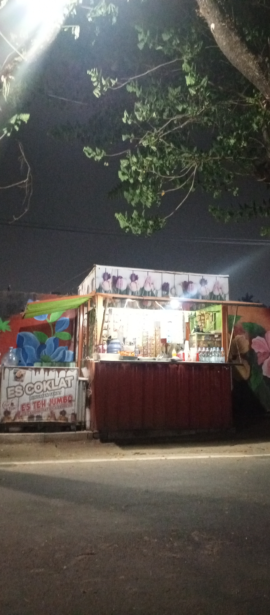 This is a night-time photograph of a food and drink stall, likely located in the city of Madiun, Indonesia, judging by the text on the signage. The stall is constructed from a shipping container, painted a dark red on its lower half. The stall has an open front, revealing a brightly lit interior where goods are displayed on shelves. These goods appear to include bottled drinks and packaged snacks. A blue water dispenser is visible on the counter. Above the counter, a sign with an illustration of tulips decorates the top section of the container. To the left of the main stall structure, there is an additional section with a green awning and a painted mural featuring large blue abstract shapes and stylized green foliage. A sign on this section reads "ES COKLAT MINUMAN SEHAT ES TEH JUMBO" in white lettering. The stall is situated next to a paved road, with a tree branch, sparsely leaved, extending into the frame from the upper left. The background is dark, indicating nighttime, with a faint glow from the stall's lighting. No people are visible in the photograph.