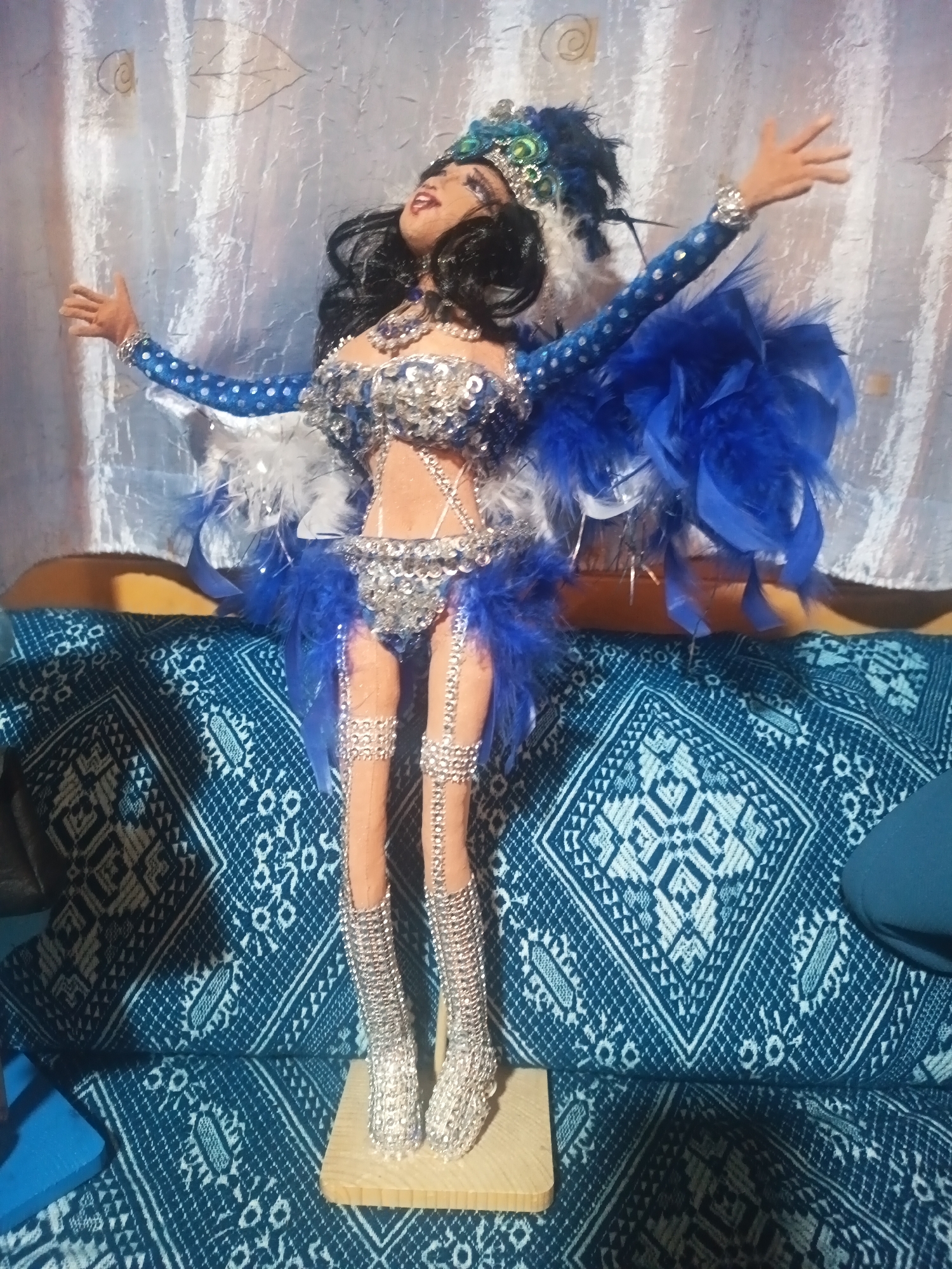 A doll in a blue and silver carnival costume stands on a wooden base, posed with arms outstretched. The background consists of a blue and white patterned fabric. The location is identified as Silistra, Bulgaria.