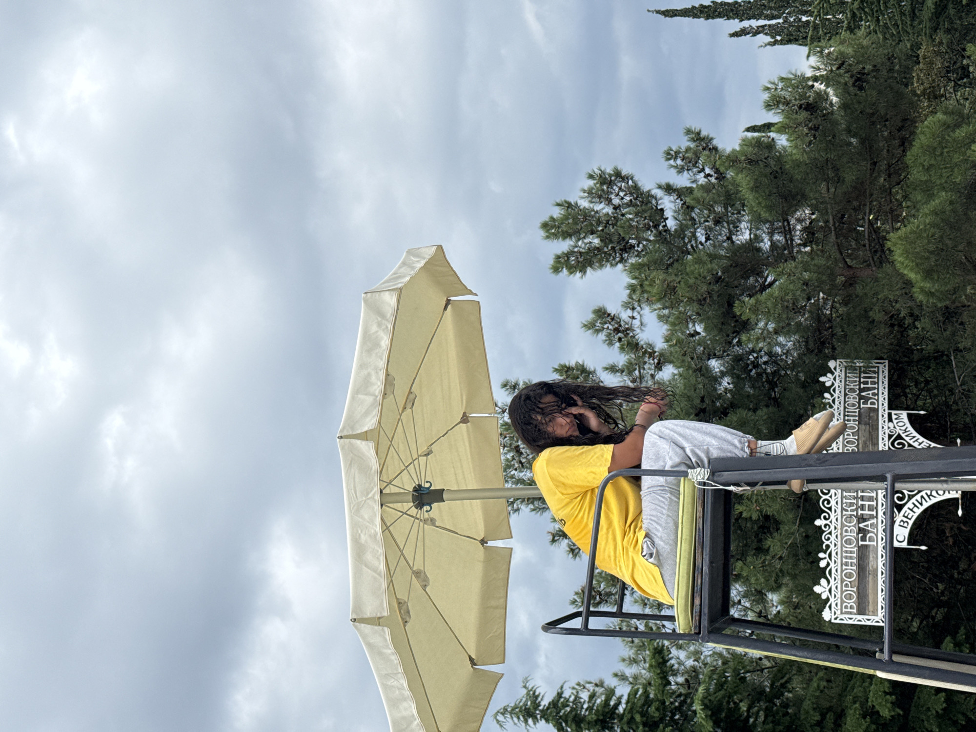A person is sitting on a high chair under a large umbrella. The person is wearing a yellow t-shirt and grey sweatpants. Their head is bowed and they appear to be tired or distressed, with their hand to their face. The chair is black and has a green cushion. In the background, there are trees and a cloudy sky. Two signs are visible below the chair with Cyrillic text that reads "ВОРОНЦОВСКИЕ БАНИ". This suggests the location is the Vorontsov Baths in Yalta. The weather appears overcast and the overall atmosphere is somber.