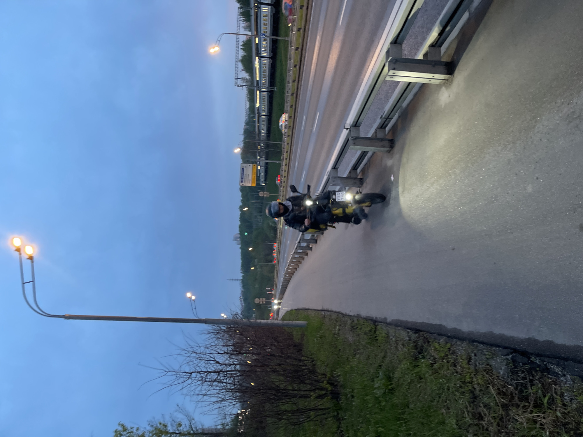 The image shows a motorcycle rider stopped on a sidewalk beside a highway in Khimki, Russia. The rider, wearing a helmet and dark clothing, is on a motorcycle with its headlight illuminating the pavement. A multi-lane highway with a train on the far side stretches into the distance, alongside a sidewalk lined with streetlights and a metal guardrail. Vegetation is visible along the roadside. The sky is overcast.