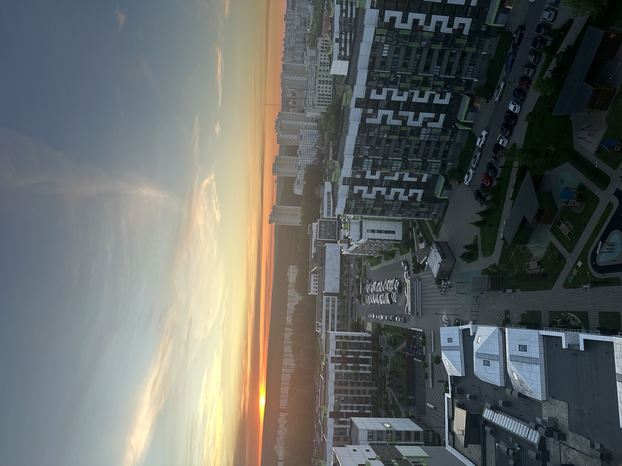 The image shows an aerial view of a city, possibly Бараўлянскі сельскі Савет, Belarus, during sunset or sunrise. The sky transitions from blue to orange and yellow, indicating the sun's position on the horizon. Tall residential buildings, some with distinctive architectural designs, dominate the right side of the image. Some buildings feature green accents. Parking lots with numerous cars are visible between the buildings. Green spaces, including trees and small parks, are interspersed throughout the urban landscape. The left side of the image shows more dense buildings along the horizon.
