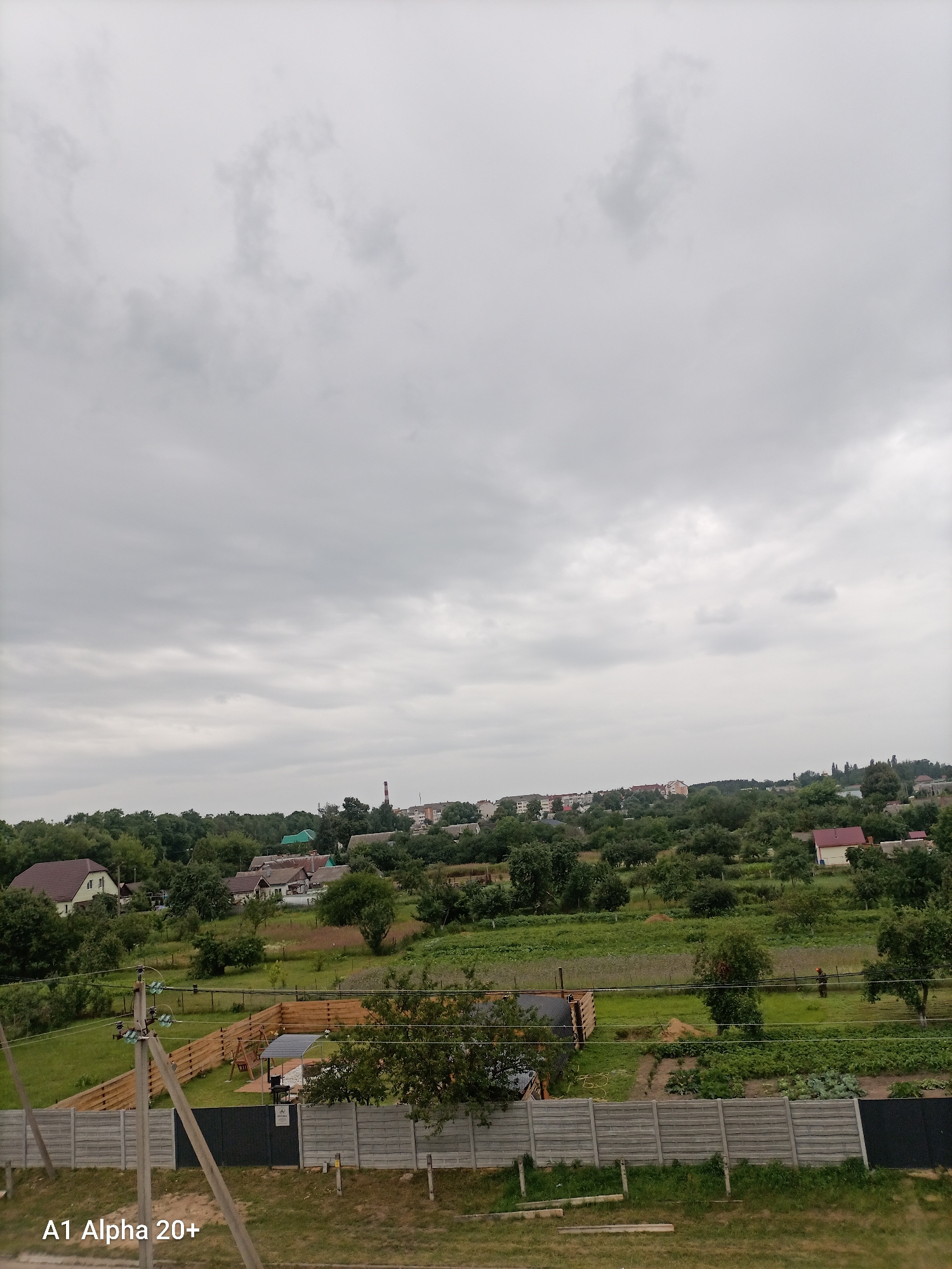 The image depicts a rural landscape under an overcast sky, likely in the Улукаўскі сельскі Савет, Belarus. The foreground features a gray concrete fence and a utility pole with wires. Behind the fence, a wooden fence encloses a grassy yard with a small structure, a barbeque grill, and a covered seating area. Further back, a field with crops is visible, and a person wearing an orange uniform is working in the field. Beyond the fields, a cluster of houses and trees form a village-like setting, with a tall red and white smokestack visible in the distance. The overall scene conveys a tranquil, everyday rural environment.