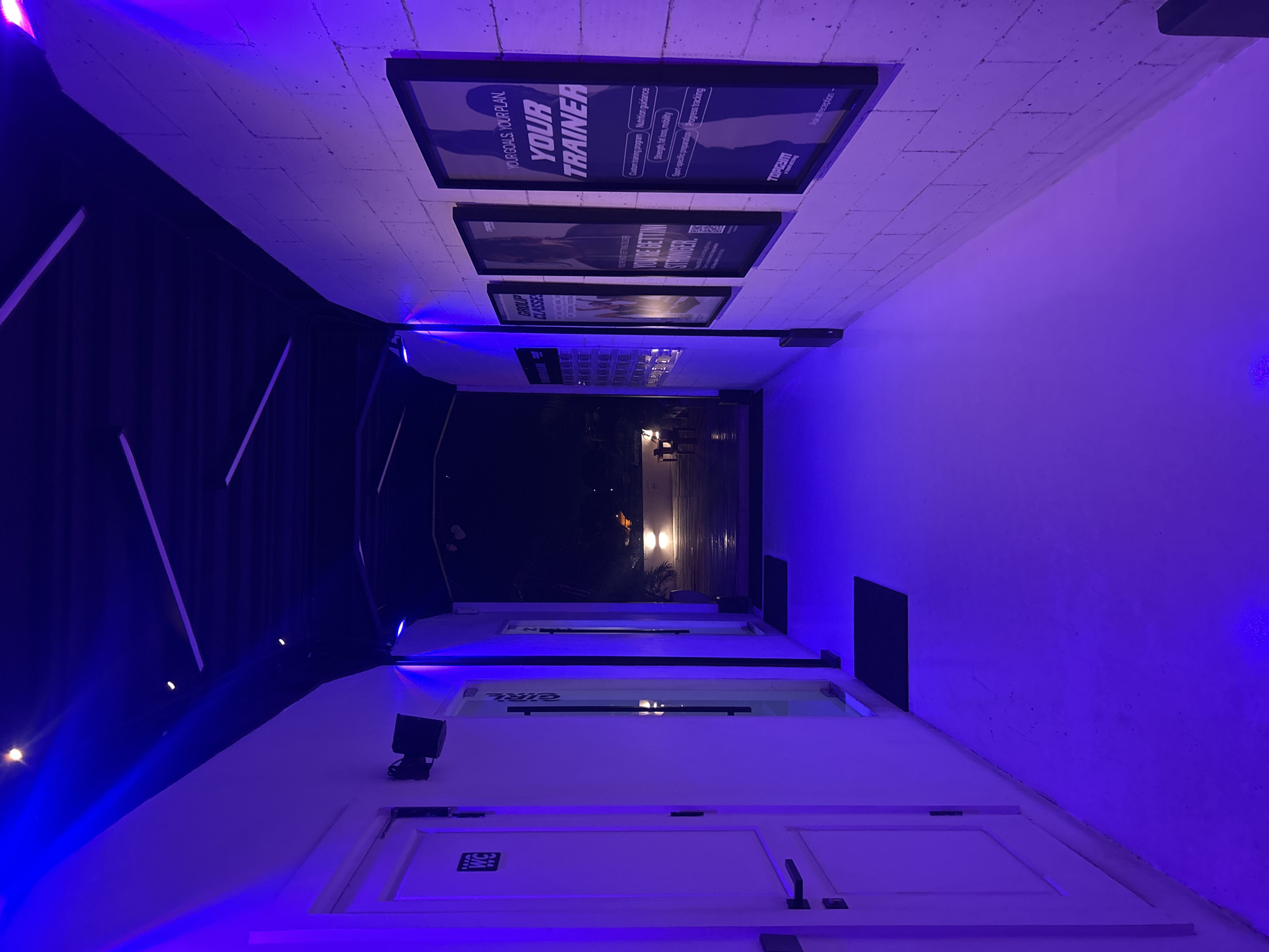 The image depicts a dimly lit hallway with vibrant purple and blue lighting, likely at a gym or fitness center in North Kuta, Indonesia. The hallway has white walls on the left, with several doors, one clearly marked with a "WC" symbol. On the right, the wall is constructed of white brick and displays several framed posters. The posters advertise fitness classes and personal training services, with text such as "GROUP CLASSES" and "YOUR GOALS. YOUR PLAN. YOUR TRAINER." The floor is a smooth, light-colored material, reflecting the colored lighting. At the end of the hallway, an open doorway reveals a glimpse of an outdoor area with wooden decking and some seating, under a night sky. The ceiling is dark and features exposed linear lighting fixtures. There are no people visible in the image.