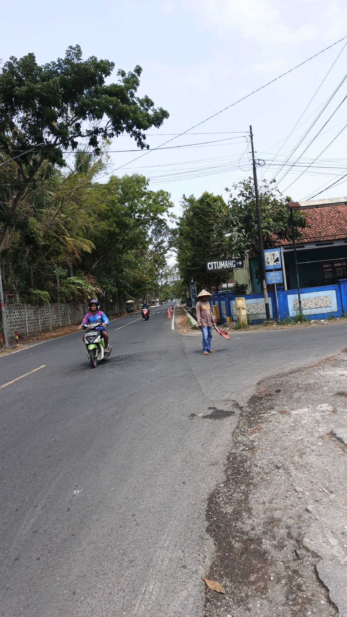 In the city of Cibenda, Indonesia, on a clear day, a person wearing a conical hat and blue jeans is standing on the right side of the road, holding a red broom. A motorcycle carrying a person wearing a blue shirt and helmet is riding on the left side of the road. The road is paved and has a yellow line marking the center. Trees line the left side of the road, and a fence is visible behind them. On the right side of the road, a building with a red-tiled roof and blue trim is visible, along with a sign that reads "CITUMANG". There are also traffic cones on the road. The sky is clear and blue.