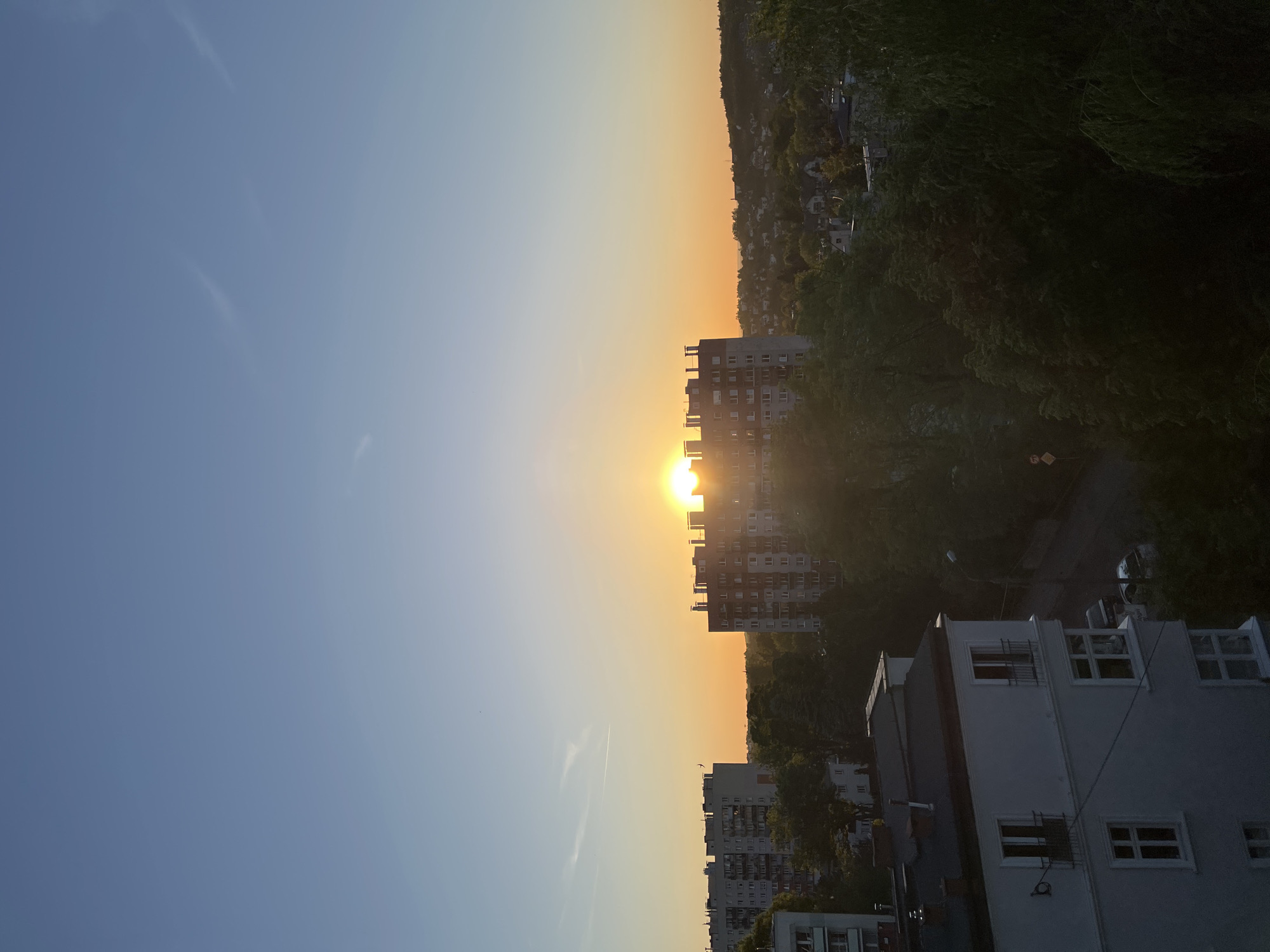 The image depicts a cityscape at sunset. The sun is positioned behind a tall apartment building, creating a bright glare. To the left of the building, the sky transitions from blue to orange. In the background, there is a tree-covered hill with houses dotted across its surface. In the foreground, there is a building. The image is taken in Unknown, Unknown.