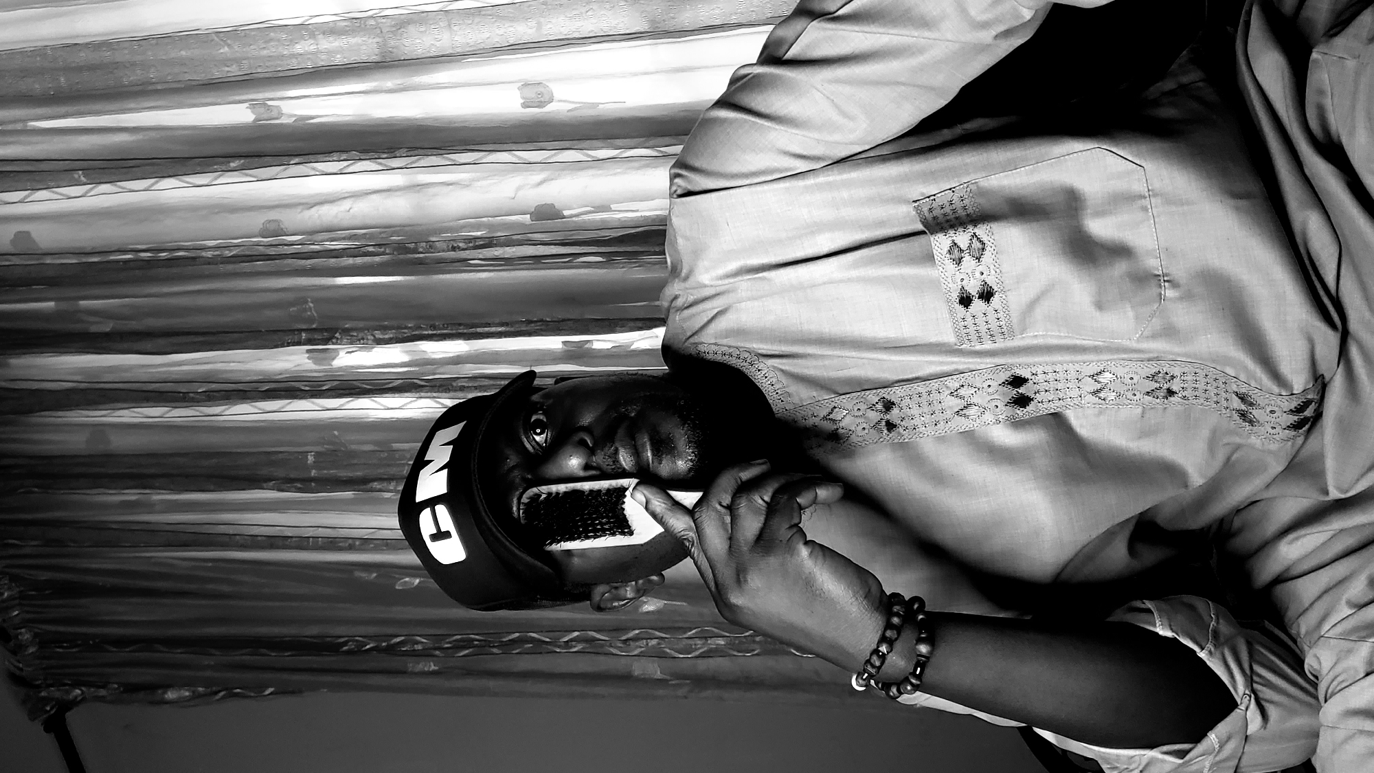 A man wearing a "GM" cap and a beaded bracelet brushes his teeth with a white-handled brush. He is wearing a light-colored shirt with decorative stitching around the neck and pocket. The background is a striped curtain. This image may have been taken indoors in Sabon Tasha, Nigeria.