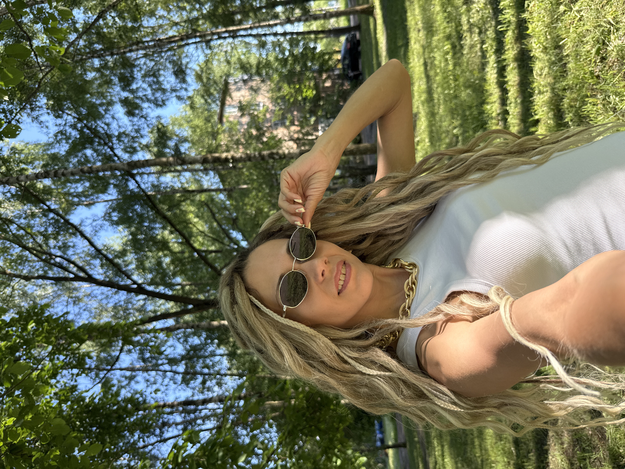 A woman with long, blonde dreadlocks and a gold chain necklace smiles at the camera while holding her sunglasses. She is wearing a white, ribbed tank top. The woman is lying down outdoors, with her arm extended and propped up on the ground. Her face is partially obscured by her sunglasses, but her smile is visible. The background is a blurred image of trees and grass, suggesting an outdoor setting. The lighting indicates it is daytime, likely sunny. The image has a casual and relaxed feel.