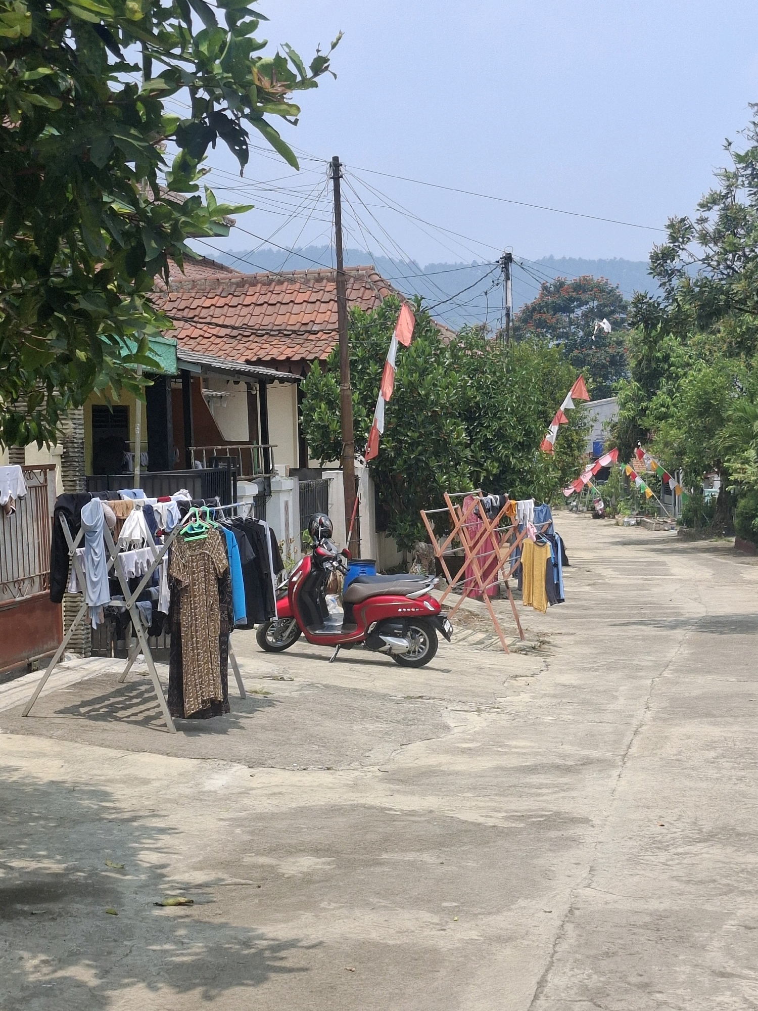 This is a medium-angle, outdoor, daytime shot of a street scene in Ciheulang Tonggoh, Indonesia, likely during a celebration given the numerous red and white flags lining the street and adorning poles. In the foreground, a clothesline holds various garments drying in the sun. To the right of the clothesline, a red scooter is parked on the concrete road. Behind the clothesline and scooter, a residential building with a tiled roof is visible, with trees and lush green foliage surrounding it. The street itself is paved with concrete and shows some signs of wear. In the background, a network of utility poles and wires crisscrosses the sky, with a hazy view of hills in the distance. The overall impression is that of a typical residential street in a tropical or subtropical climate, with a festive atmosphere.