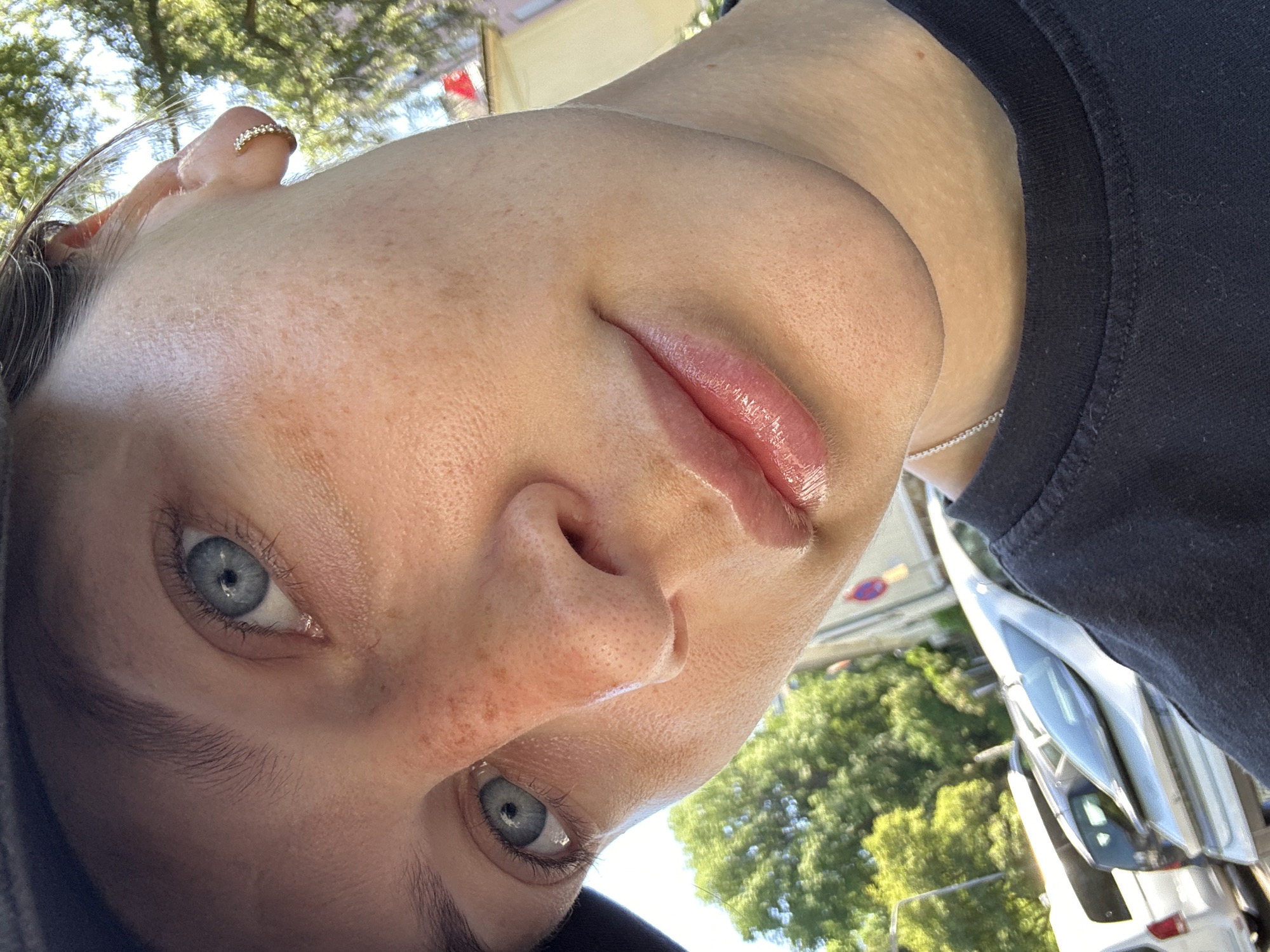 A close-up, tilted selfie shows a fair-skinned person with freckles and light blue eyes. They have a nose ring with small jewels. They are wearing a black t-shirt and a silver necklace. The background is blurred, but contains trees, cars, and signage. Due to lack of specific information in the background, I cannot confirm that this image was taken in the city of Dillfeld, Germany.