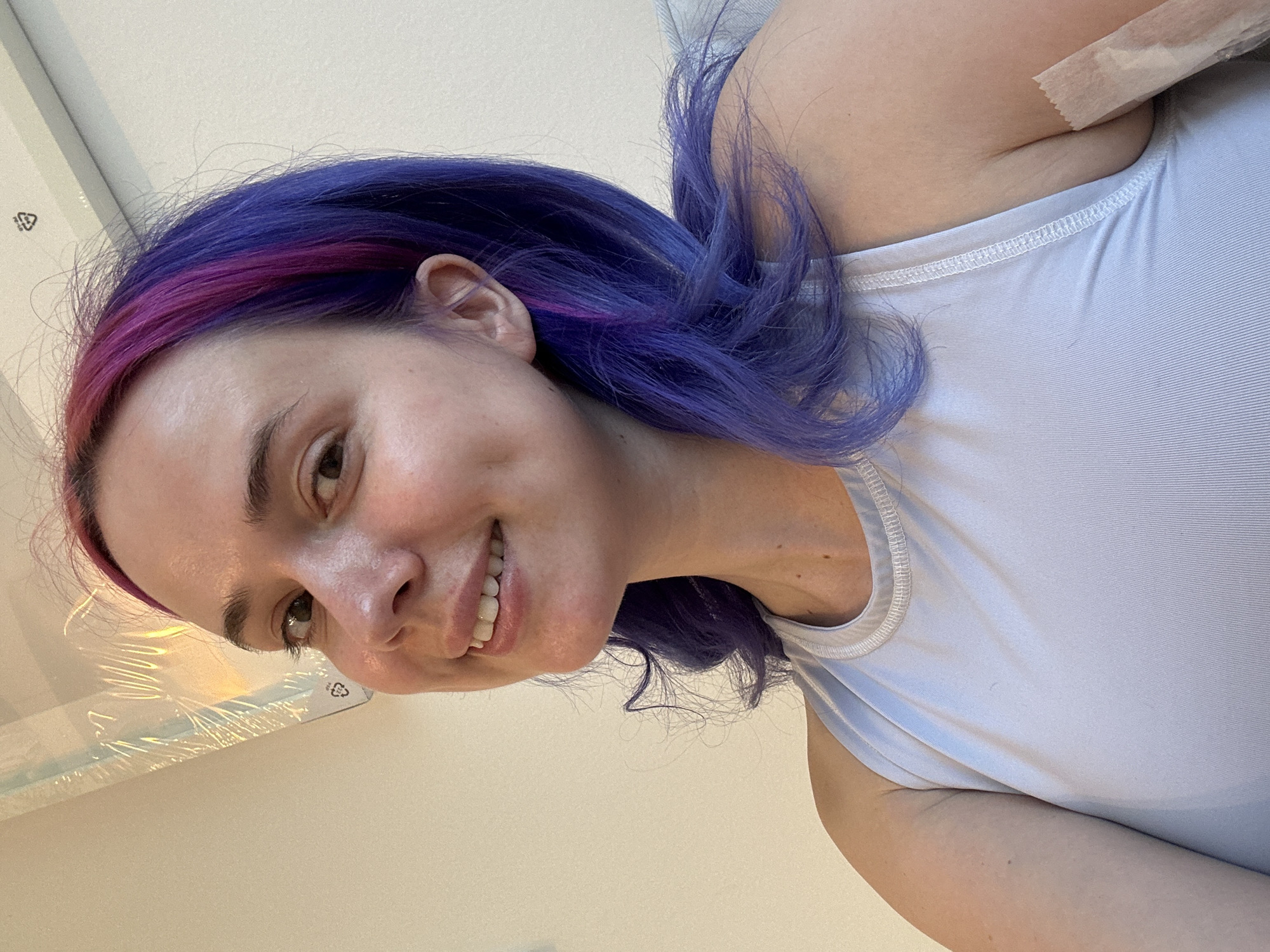 A woman with vibrant, multi-toned hair, primarily purple with streaks of magenta, is featured in a close-up, upward-facing selfie. Her head is tilted, and the image is rotated, suggesting a dynamic, casual pose. She has fair skin, a wide, genuine smile revealing white teeth, and bright eyes. Her eyebrows are dark and well-defined.
She is wearing a light blue or lilac-colored ribbed tank top. Visible near her right shoulder/collarbone area is a piece of light-colored medical tape or similar adhesive, slightly crinkled. Her hair is styled loosely, with some strands falling around her face and shoulder.
The background is simple and light-colored, likely a wall or ceiling, indicating an indoor setting. In the upper left corner, behind her head, a part of a plastic-wrapped object is visible. On this object, a black recycling symbol (a triangle made of three arrows) is clearly discernible. The lighting appears natural and bright, suggesting it is daytime. The woman's expression conveys happiness and friendliness. The scene takes place in the City of Zagreb, Croatia.