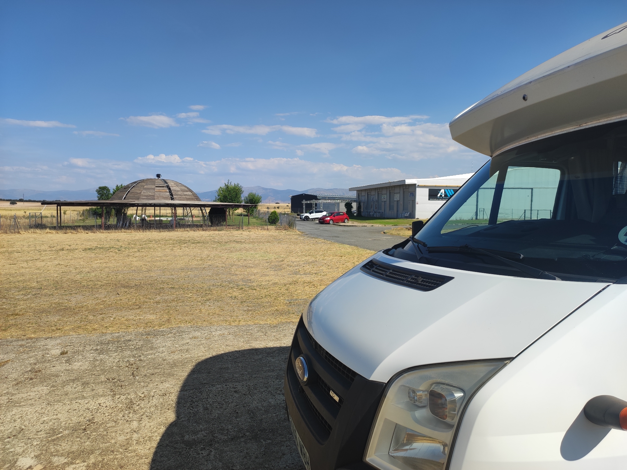 The image, captured in Marugan, Spain, depicts an outdoor scene with a prominent white camper van in the foreground, angled towards the right. Behind the van, a dry, golden field stretches towards the horizon. In the mid-ground, a circular wooden structure, possibly a pavilion or shade, stands in the field. Further back, several buildings with dark roofs and some lighter structures are visible, including a building with signage that reads "A ESCUELA DE PILOTOS." Two cars, one white and one red, are parked in front of one of the buildings. The sky above is a clear blue with scattered white clouds, and distant mountains are visible on the horizon. The overall environment suggests a rural or semi-rural location.