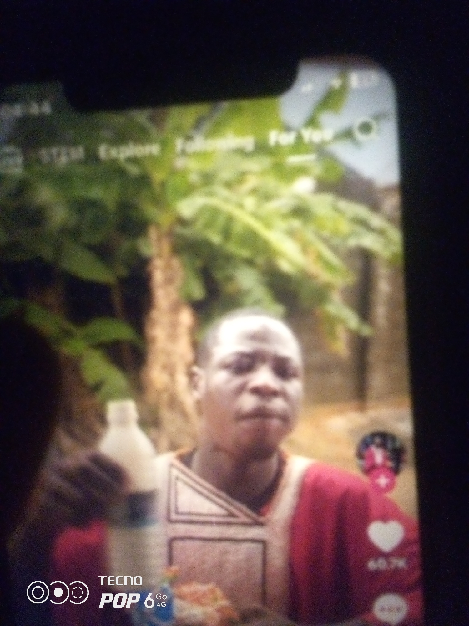 The image depicts a close-up shot of a young male, likely of Nigerian descent, viewed through a mobile phone screen. He is wearing a red garment with white trim around the neckline and chest area. In his left hand, he holds a white bottle. The background is a blur of green foliage, suggesting an outdoor setting with trees and possibly a banana tree. The overall scene appears to be casual and possibly a captured moment from a video. There is no discernible indication of the city of Howayi, Nigeria, within the image itself. The image is a photograph of a screen displaying content.