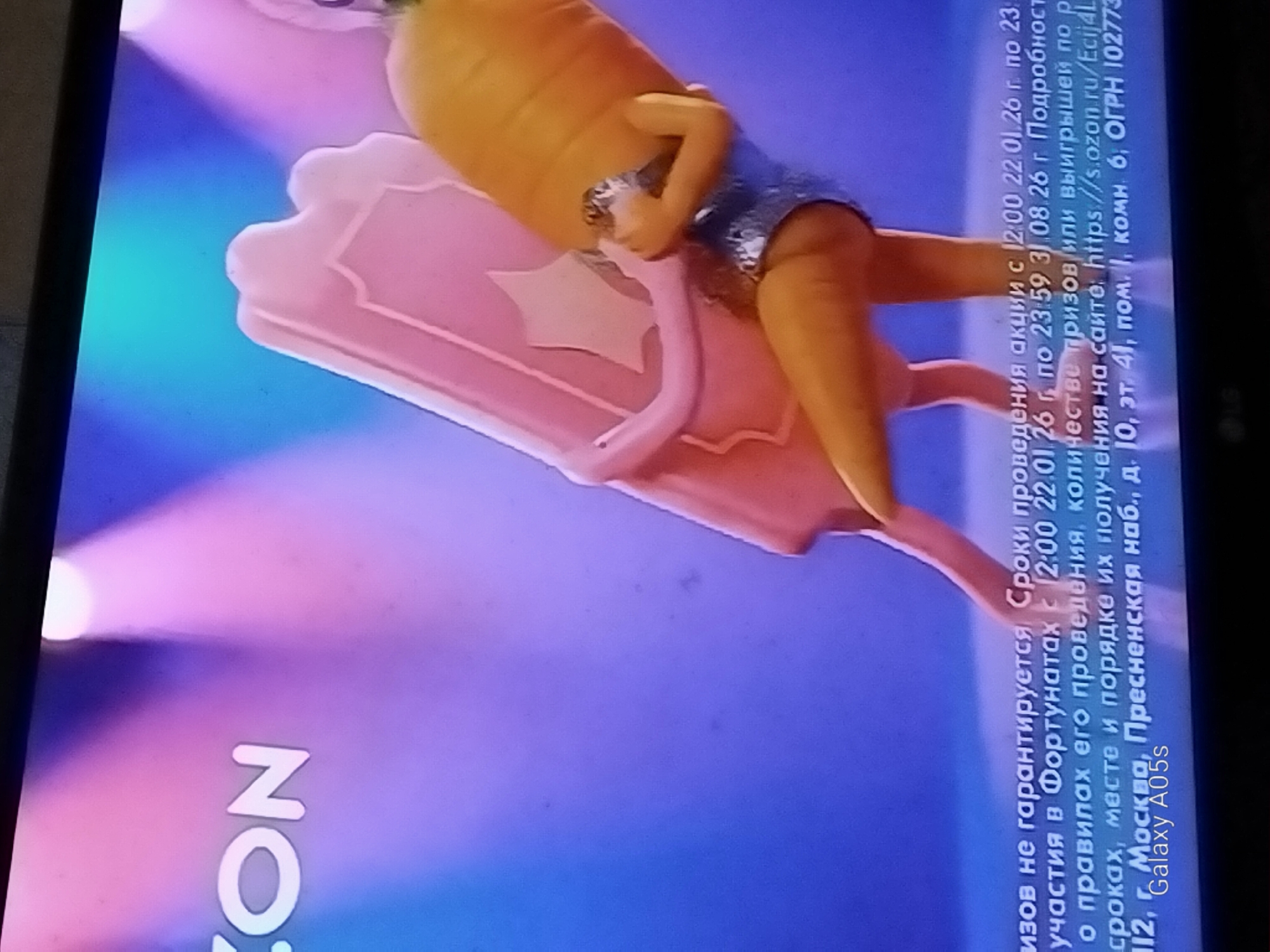 The image shows a television screen displaying a colorful advertisement. The advertisement features cartoonish characters, one of which resembles a carrot wearing a sparkly blue outfit. This character is posed as if interacting with a pink, rectangular object. The background is a gradient of blue and pink, with spotlights creating a stage-like effect. Text is visible at the bottom of the screen, providing promotional details, including dates, a website address (https://s.ozon.ru/Ecij4L), and an OGRN number (1027739321744). The text also mentions "Galaxy A05s," likely indicating a promotion related to this phone model. The text mentions "Москва, Пресненская наб., д. 10, эт. 41, пом. 1, комн. 6," which refers to a location in Moscow, Russia, not Druzhba, Russia. There is no information or visual element in the image that links it to the city of Druzhba, Russia.
