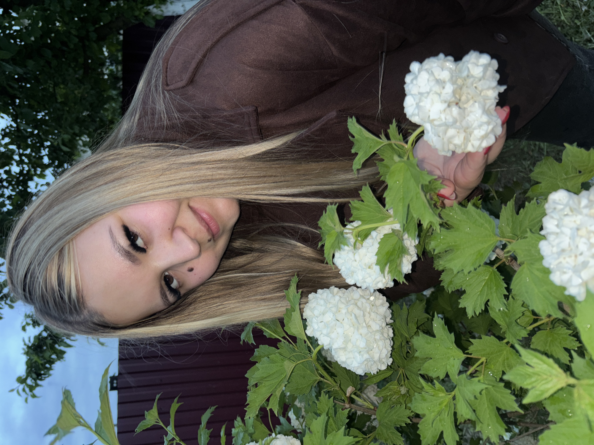 A woman with long blonde hair wears a brown jacket and is holding a white flower in her right hand. She is standing in front of a bush with white flowers and green leaves. In the background, there is a dark red fence and trees. The image appears to be taken outdoors, possibly in a garden or park. The location is specified as Фоки, Russia.