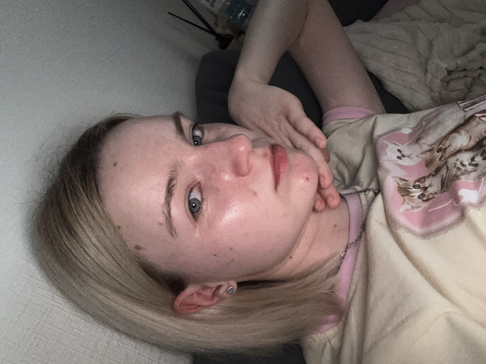 A close-up, slightly angled, indoor image features a fair-skinned person with light blonde hair. The person is lying down, with their head tilted slightly. Their right hand is resting on their cheek. They have blue eyes and red lips. They are wearing a light-colored top with a pink trim and a pattern of cats. A silver necklace is visible around their neck. The background includes a white wall to the left, a dark gray upholstered object, and a fluffy blanket. The image quality is slightly grainy, possibly due to low lighting. The location in the city of Unknown, Unknown is not apparent from the visual information.