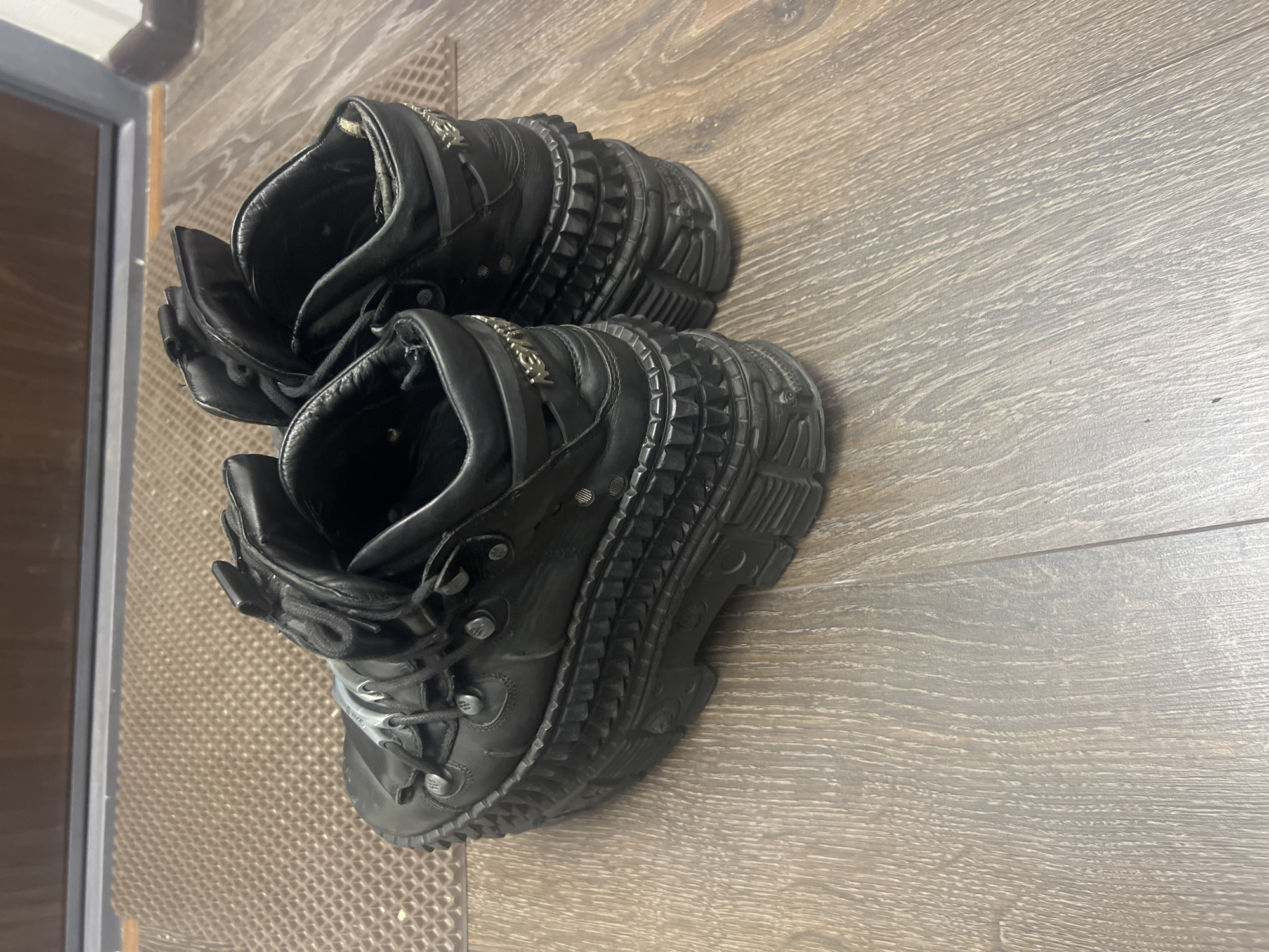 Two black, heavy-duty platform shoes are prominently featured, lying on a light-colored wooden floor. The shoes appear to be made of black leather or a similar material, with thick, multi-layered soles that exhibit a textured, ridged, and somewhat spiked design. Both shoes are laced up, and the lacing system includes metal eyelets or hooks.
Visible details on the shoes include metallic emblems on the sides of the upper part, which clearly spell out "NEW ROCK". There are also smaller, stylized symbols or letters, possibly "NR," on some of the metal lace fasteners. The shoes are positioned diagonally across the frame, with the toes pointing towards the upper right and the heels towards the lower left.
The setting is indoors, with the shoes resting on what appears to be laminate or hardwood flooring characterized by a light brown, greyish tone with visible wood grain. To the left side of the image, part of a darker, possibly wooden or metallic, door or window frame is visible, along with a textured, lighter-colored mat or floor covering. There is no activity taking place, and no people are present. The lighting suggests an indoor environment, either from artificial sources or indirect natural light, making the time of day and weather unidentifiable.