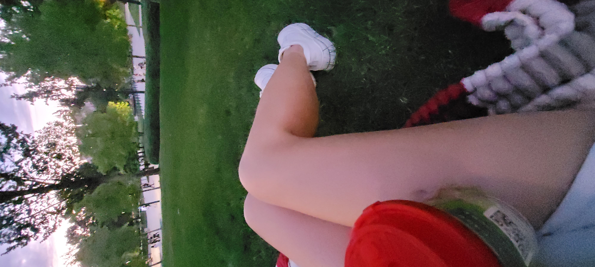 In an outdoor park setting, a person's legs are visible, sitting on a grassy area. The person is wearing white sneakers. In the foreground, a red cup is held by the person's left hand. The background features trees, a pathway, and people walking in the distance. The sky is visible, with a hint of twilight. The overall lighting suggests it is late afternoon or early evening.