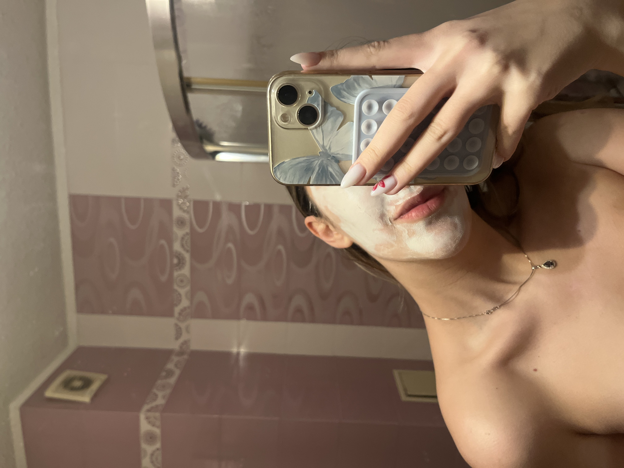 A person takes a selfie in a bathroom. The person has a face mask on and is holding a smartphone with a flower-patterned case. The person's manicured hand is visible. The bathroom has pink and white patterned tiles. A towel rack is visible on the left side of the frame. The city of Sofia, Bulgaria, cannot be determined.