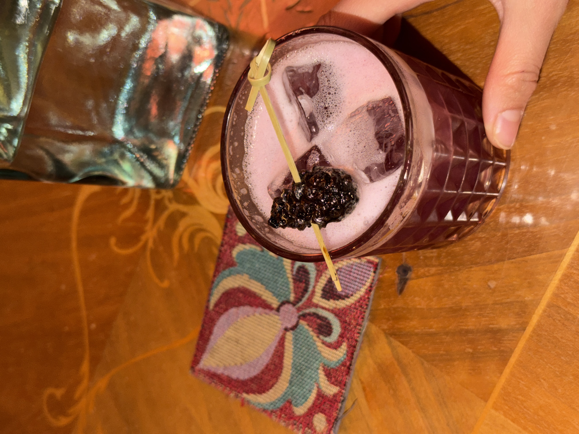 This image, captured in Krasnoyarsk, Russia, shows an overhead view of a hand holding a cocktail glass. The cocktail itself is a vibrant pink or light purple, topped with a frothy layer and containing ice cubes. A single, dark blackberry is skewered on a small bamboo pick, serving as a garnish.
The glass, which has a distinct textured or faceted pattern on its exterior, rests on a colorful fabric coaster. The coaster features an intricate, symmetrical floral or abstract design in shades of red, blue, light green, pink, and cream. The entire setup is placed on a warm-toned wooden table, which displays subtle decorative swirling patterns across its surface.
To the left of the cocktail, a highly reflective, silvery metallic object occupies a portion of the frame, possibly a bag or part of another container. A small, dark, key-shaped item is also visible on the wooden table, slightly to the right and below the glass. The scene depicts someone enjoying or about to enjoy a beverage in what appears to be an indoor setting. No discernible text is visible in the image.