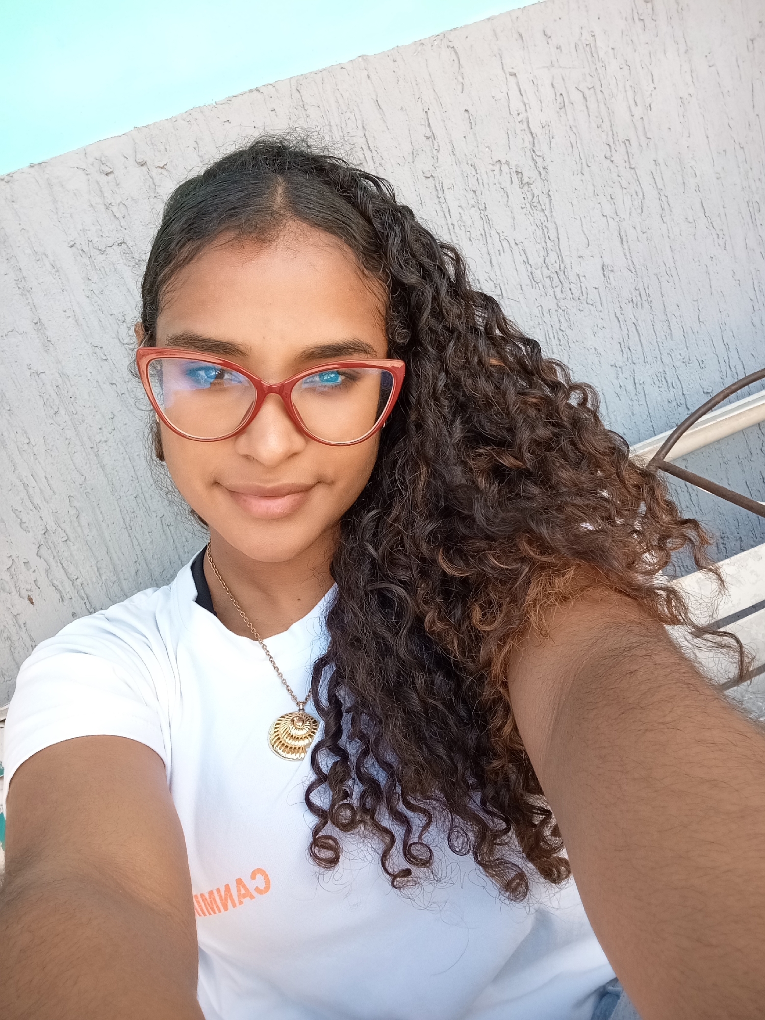 A young woman takes a selfie outdoors. She is wearing red glasses and a white t-shirt with orange writing. She has long, curly hair and a gold necklace. The background is a textured gray wall and a blue sky. The photo was taken in Juan Griego, Venezuela.