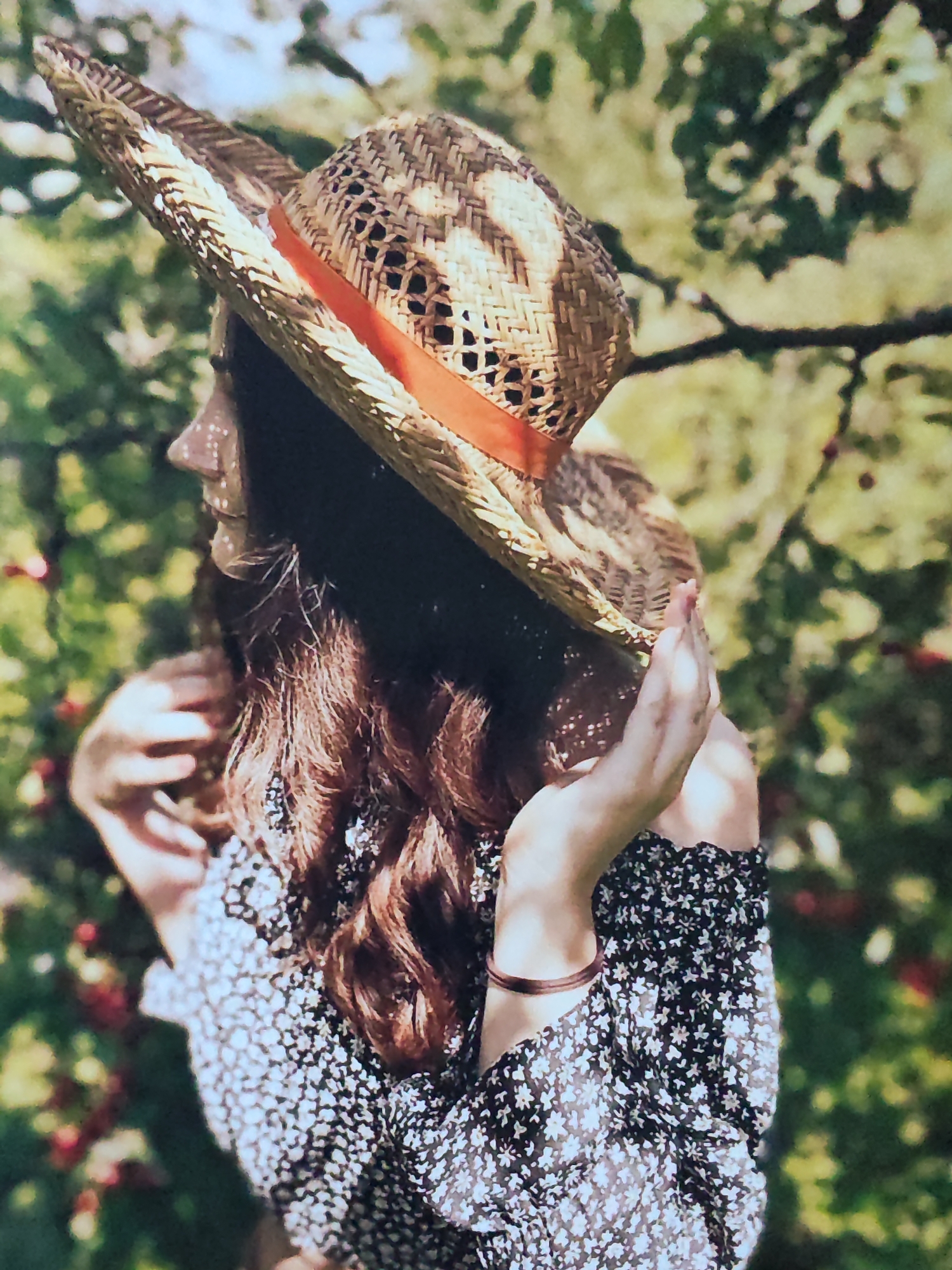 A medium shot photograph captures a person from the chest up, wearing a straw hat adorned with an orange ribbon. The person, facing to the left, has long, wavy brown hair that extends past their shoulders. They are also wearing a black off-the-shoulder dress with a white floral pattern. Their right hand is raised, touching their chin, and their left hand is near their chest. The background is softly focused, showing lush green foliage and hints of red berries, suggesting an outdoor setting, possibly a garden or natural landscape. The lighting appears to be natural sunlight, casting dappled shadows on the person's face and dress.