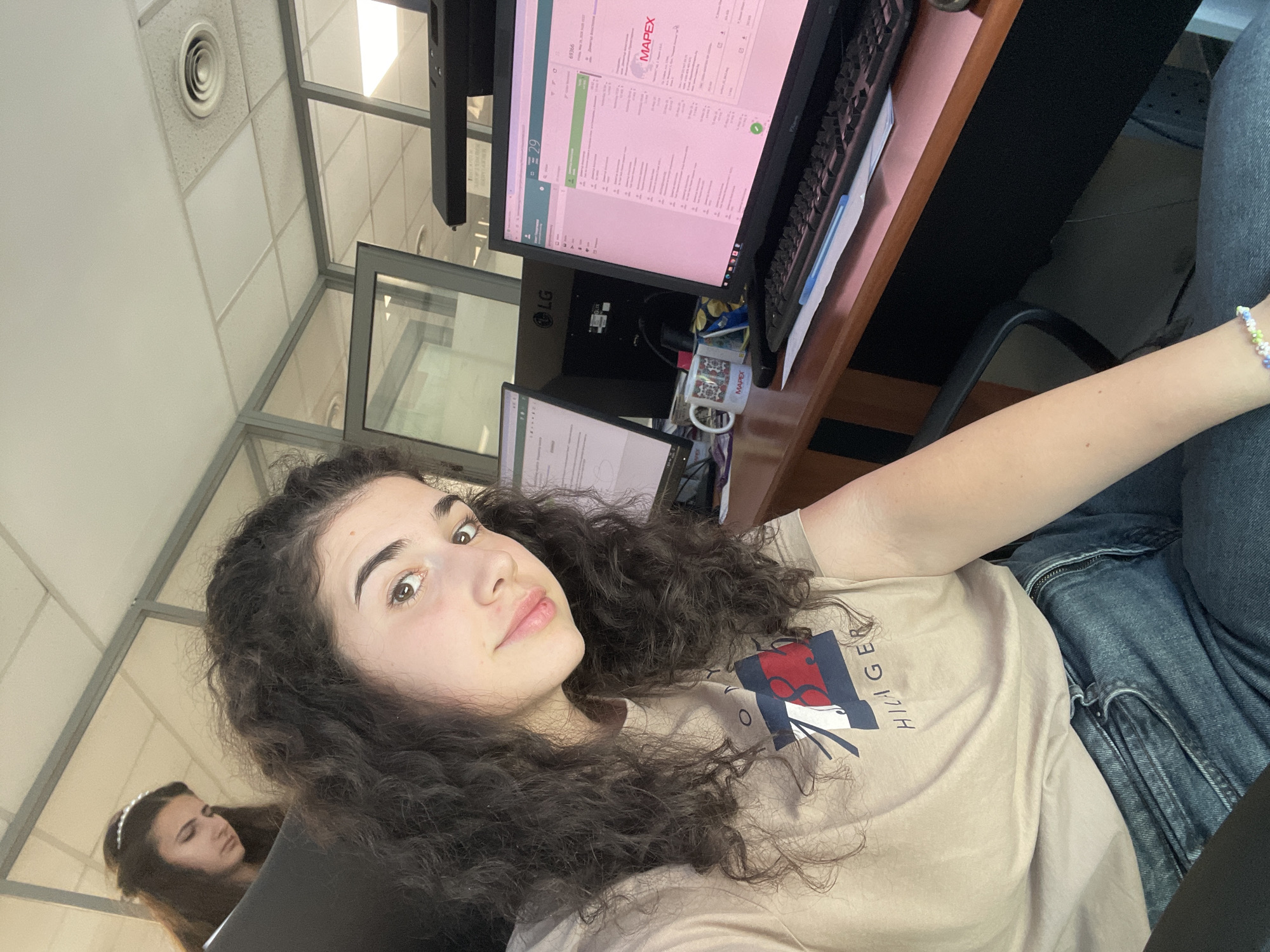 A young woman with curly dark hair is sitting at a desk in an office. She is wearing a light brown t-shirt with a red and blue logo and blue jeans. She is smiling slightly at the camera. In the background, there are computer monitors, a keyboard, and office cubicle partitions. There is another young woman with dark hair sitting in the background near the partitions. A mug with "MAPEX" printed on it sits on the desk, indicating a possible company affiliation. The office appears to be well-lit with a drop ceiling with air vents. The location is likely Sofia, Bulgaria, based on the information on the screen (MAPEX).