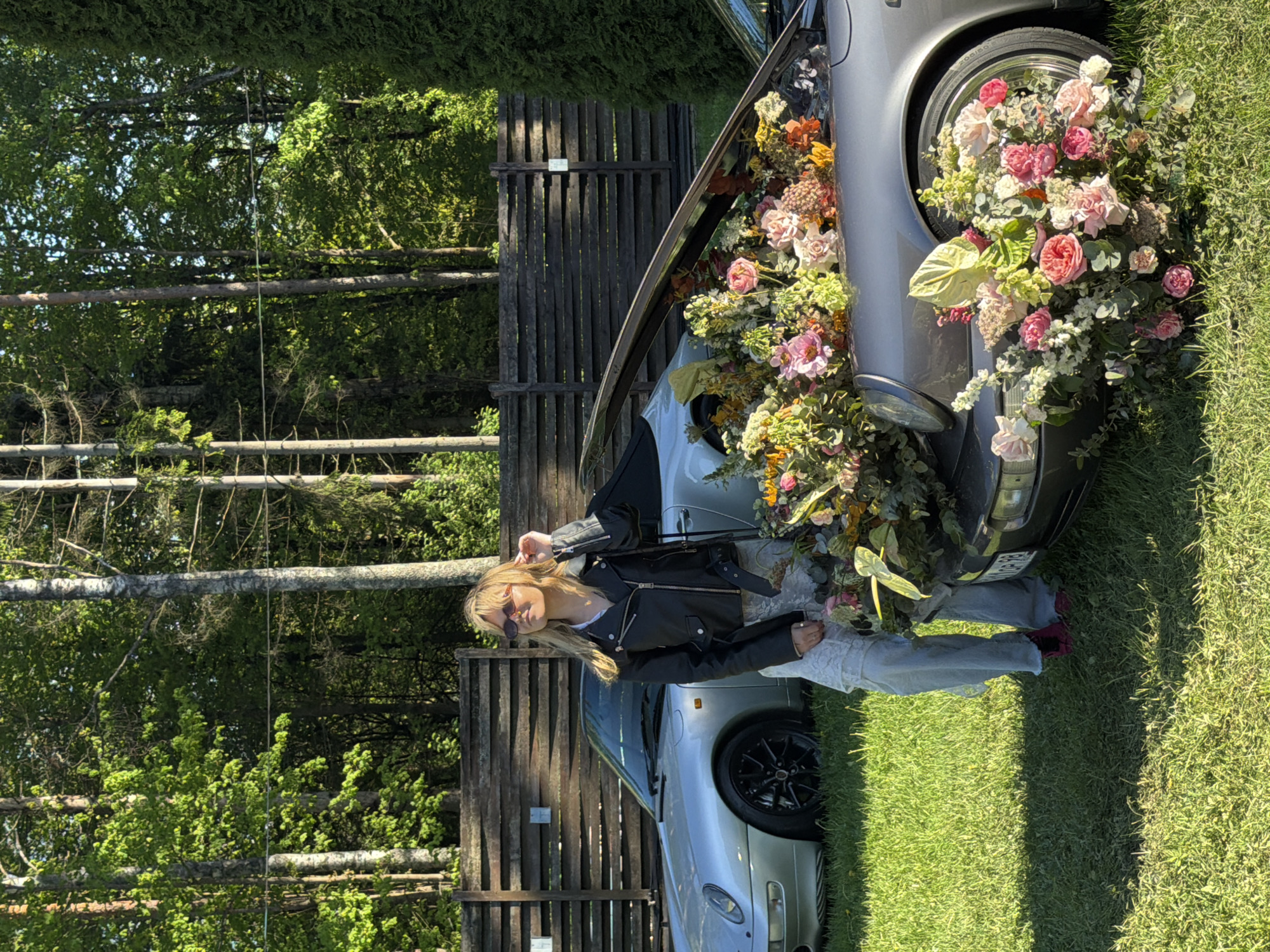 The image shows a young woman standing next to a silver Porsche car. The car's front is filled with an arrangement of flowers and greenery, extending from the open hood and encompassing the front tire. The woman is wearing a black leather jacket, a white top, light-colored pants, and sunglasses. She is touching her hair with her right hand. The background features a wooden fence, trees, and green grass. The scene is taking place outdoors on a sunny day. The license plate on the car indicates that the location is in Repino, Russia.