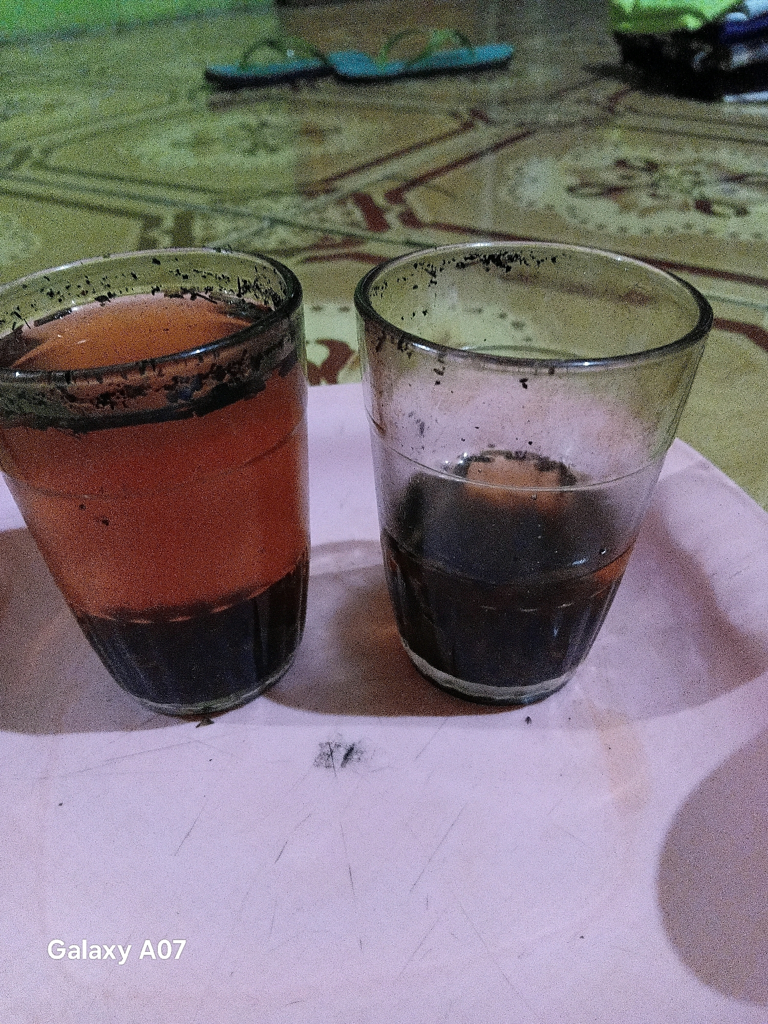 Two glasses of dark liquid, likely coffee or tea, sit on a pink tray. The glass on the left appears to have more of a reddish-brown hue, while the glass on the right is a darker, almost black color. Both glasses have remnants of the beverage clinging to the rims and the sides. The tray itself has some smudges and visible textures. In the background, a patterned surface, possibly a rug or mat, is visible, with a blurred green element at the very top. Further back, a pair of blue flip-flops are faintly discernible. The overall lighting suggests an indoor setting, possibly at night or in a dimly lit room. The bottom left corner of the image displays the text "Galaxy A07", indicating the device used for capturing the photograph. The scene is located in Margo Rukun, Indonesia.