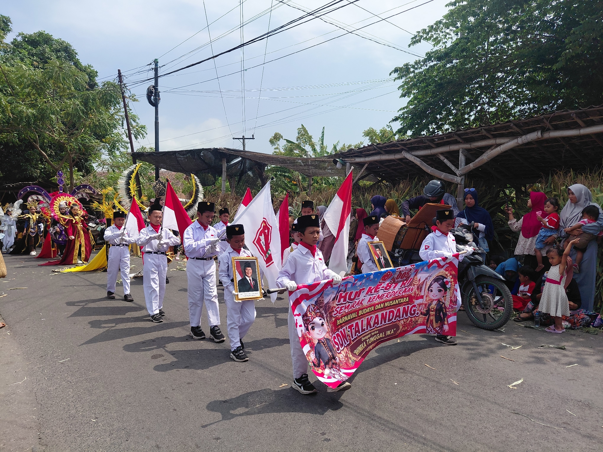 In the city of Bimo, Indonesia, a parade is underway on a sunny day, with a clear blue sky and scattered clouds overhead. The street is paved and appears to be lined with trees and some simple, covered structures. On the right side of the image, people are gathered, some sitting on the ground, observing the procession. Among them are women in headscarves, children, and a person on a motorcycle.
The parade features a group of children dressed in white uniforms with red scarves and black hats, marching in formation. They are holding Indonesian flags, and some are carrying framed portraits of an adult. A prominent banner, also in red and white, is being carried by two children. The banner displays festive illustrations and text in Indonesian, indicating a celebration for "HUT KE-81 KEMERDEKAAN REPUBLIK INDONESIA" (81st Anniversary of the Independence of the Republic of Indonesia) and mentioning "KARNAVAL BUDAYA DAN NUSANTARA SON TALKANDANG" (Cultural and Archipelago Carnival Son Talkandang).
To the left of the marching children, there is a figure in a more elaborate costume, possibly part of a different parade unit, adorned with bright colors and headdress elements. The overall scene suggests a local community event, likely a celebration of national independence.