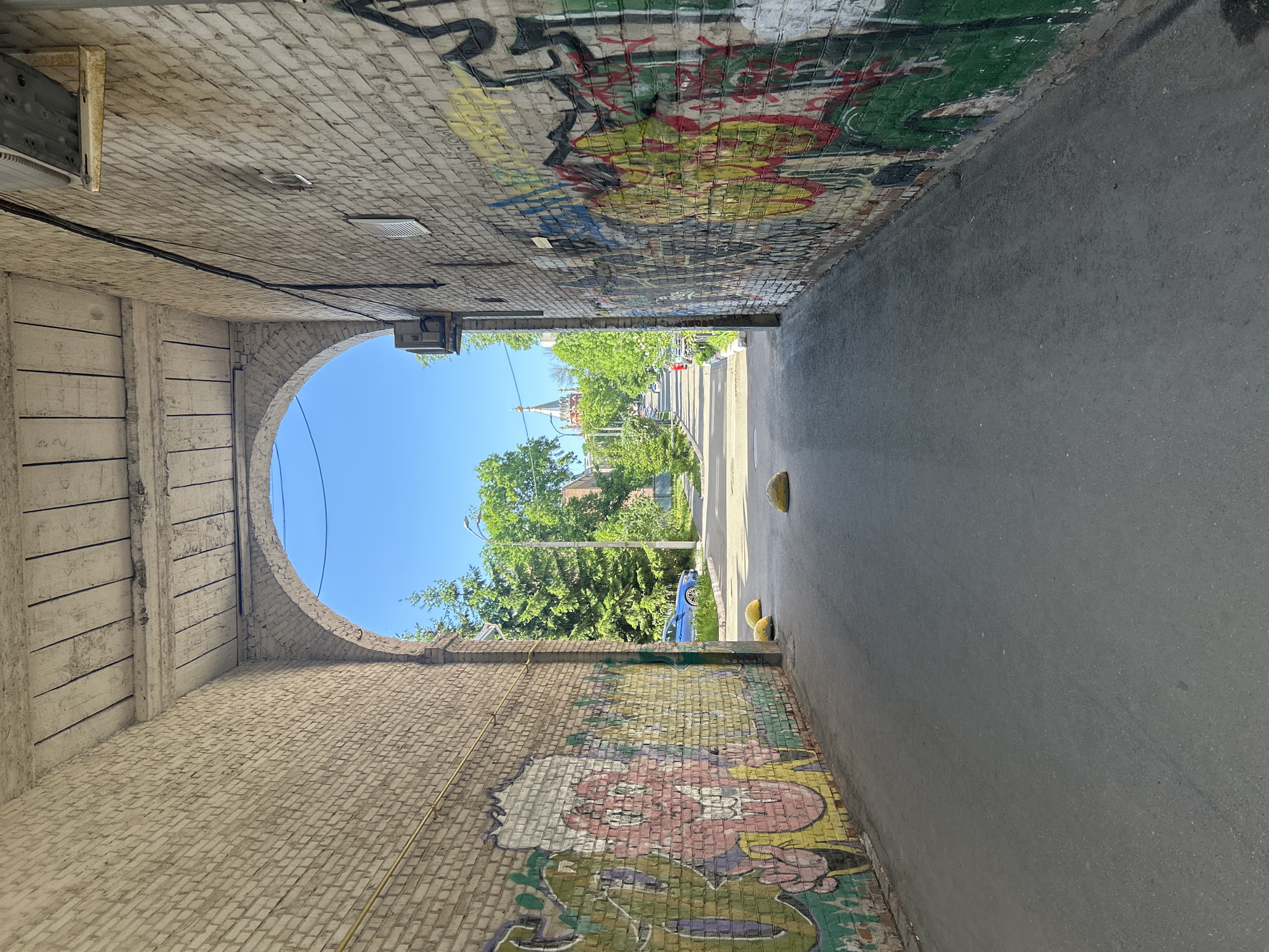 A street view of Izhevsk, Russia, through an urban underpass. The underpass walls are brick, decorated with graffiti. The left wall is aged, off-white, while the right wall features more colorful and complex graffiti. The underpass has an arched entry on the left side. Beyond the underpass is a street with greenery, including trees and grass. In the distance, a building with a prominent steeple can be seen. Three yellow bollards are located on the edge of the street. A blue car is visible parked on the side of the road. The sky is clear and blue. The road surface is asphalt.