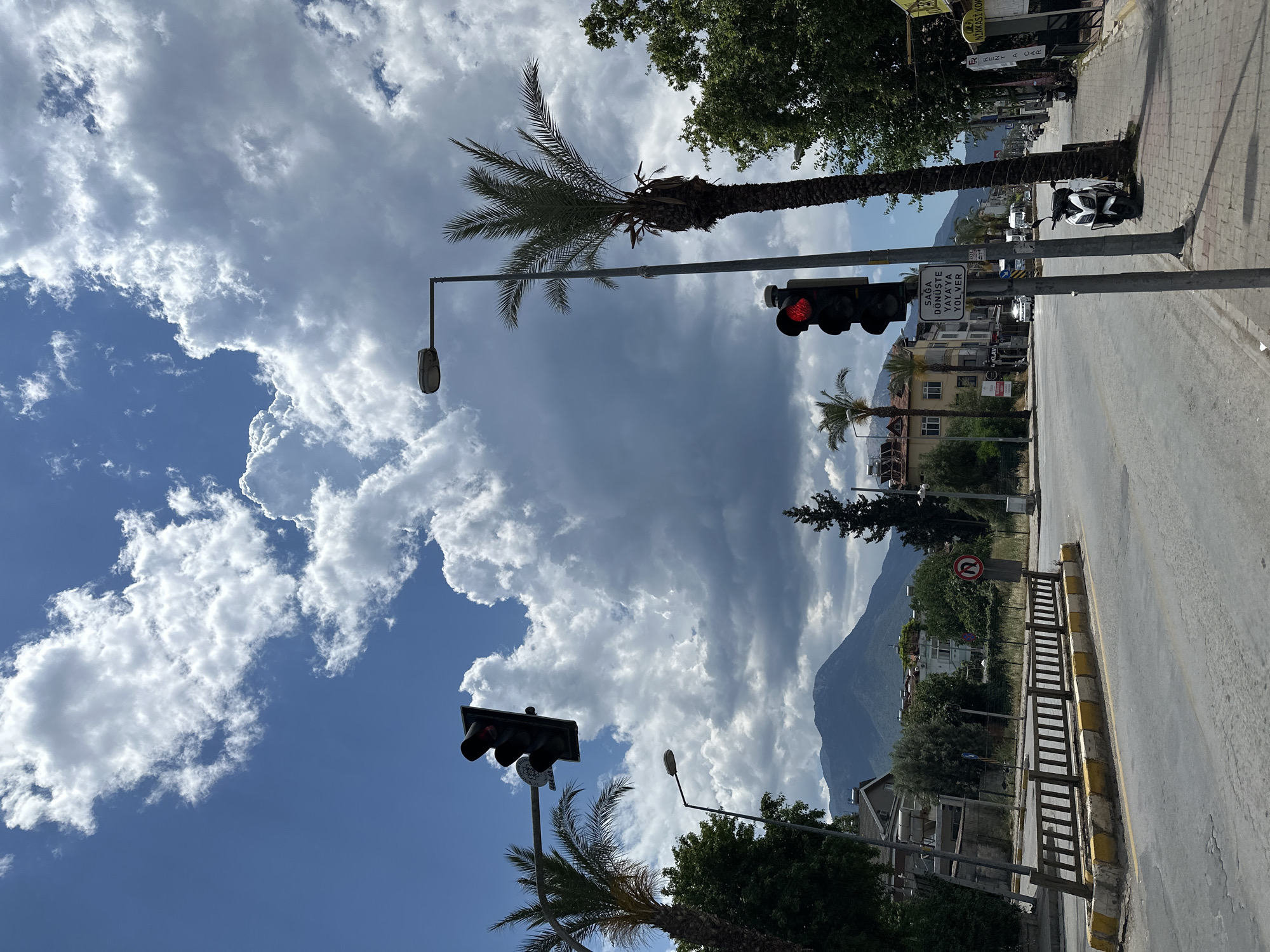 The image captures a street scene in Fethiye, Turkey. The sky is a mix of clear blue and dramatic, dark storm clouds. Palm trees line the street, some juxtaposed against traffic lights showing red. There are buildings and trees in the background. The road is empty, and a mountain looms in the distance. A scooter is parked along the sidewalk.