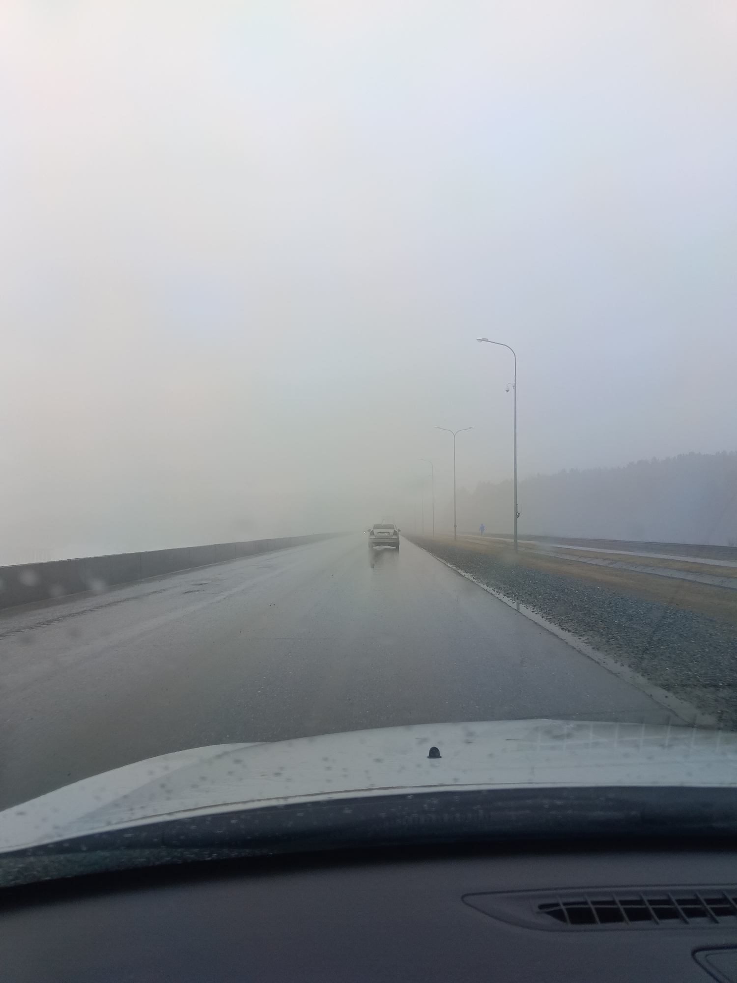 The image shows a view from inside a car driving on a wet road in Chaikovsky, Russia. The road is a bridge or causeway with concrete barriers on the left. There is light-colored vehicle in the distance, traveling in the same direction. A row of streetlights lines the right side of the road, with gravel alongside the road beyond the curb. The sky is overcast and foggy, obscuring the view. A dark line of trees or forest is visible on the right horizon. The car’s dashboard and windshield are visible in the foreground. Raindrops are visible on the windshield.