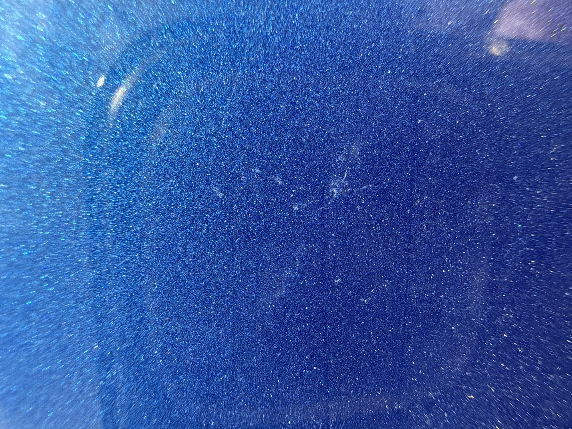 The image is a close-up, abstract shot of a sparkling blue surface. The surface is textured with numerous tiny, reflective particles that create a glittering effect, reminiscent of metallic paint or glitter. There are faint, irregular smudges or light reflections on the surface, particularly in the central and upper right portions of the image. The lighting suggests a smooth, possibly curved, material. There are no people, identifiable objects, or environmental features visible. This image is not locatable to a specific place in the city of Khmilna, Ukraine, or any other location, as it depicts a texture rather than a scene.