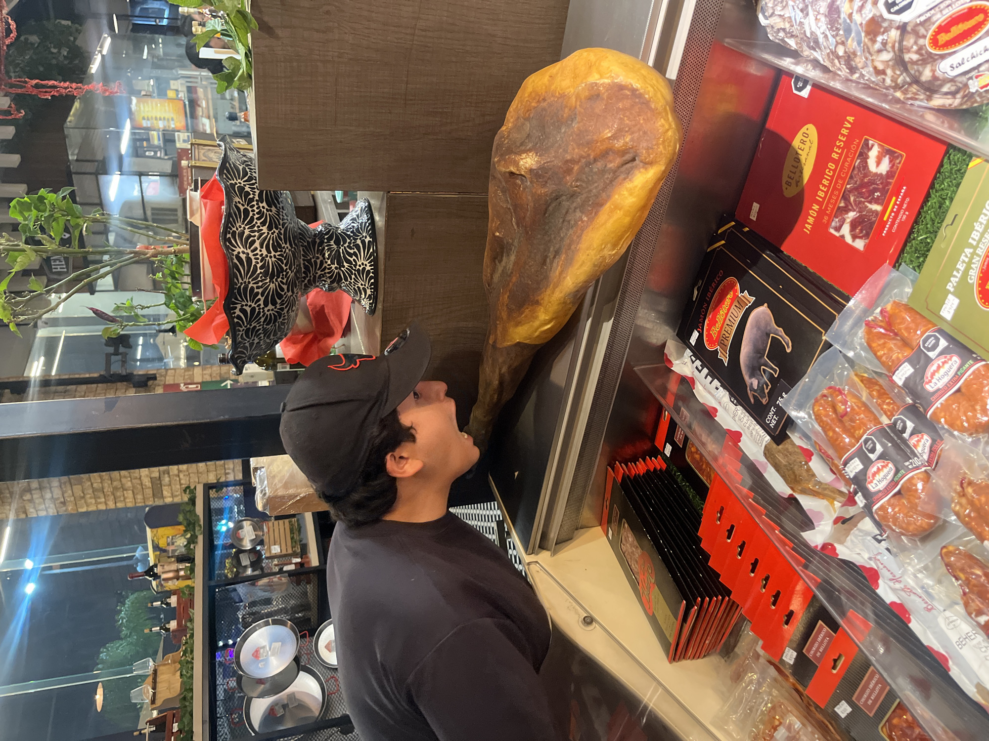 A man in a black t-shirt and a black cap with a red logo is looking upwards with his mouth open towards a large jamón leg. The jamón is located inside of a refrigerated display case, alongside various packaged meat products including "Bellotero" jamón, "La Hoguera" chorizo, and "Bellidonco" salchicha. Reflections in the glass indicate the location to be a store, with visible shelves holding wines and other goods. The location is Puebla, Mexico, as determined by the OCR identifying "Jamón Ibérico" and other Spanish-language product names.
