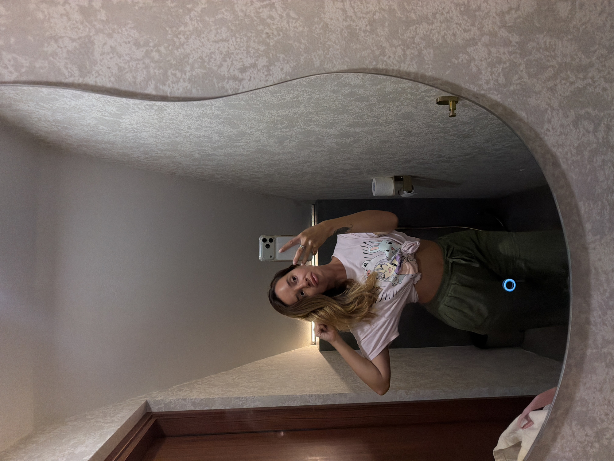 A woman is taking a mirror selfie in what appears to be a bathroom. She is wearing a light pink t-shirt with cartoon characters printed on it and olive green pants. The t-shirt is tied at the bottom, exposing a bit of her midriff. Her hair is a light brown, and she has it styled in loose waves. She is wearing a ring on her left hand.
The mirror is large and has a distinctive wavy shape at the top. The wall behind the mirror has a textured wallpaper with a light, speckled pattern. In the reflection, a toilet paper holder with toilet paper is visible on the wall, along with a small, circular blue light that appears to be a smart home device. The lighting in the room seems to be coming from an overhead fixture, casting a warm glow.
The overall scene suggests a casual, personal moment captured in a private space. The woman's pose and facial expression indicate she is comfortable and perhaps playful. The location context is Kuta Selatan, Indonesia, but there are no specific visual cues in the image to confirm this. The time of day is not discernible, nor is any information about the weather. There is no visible text in the image.
