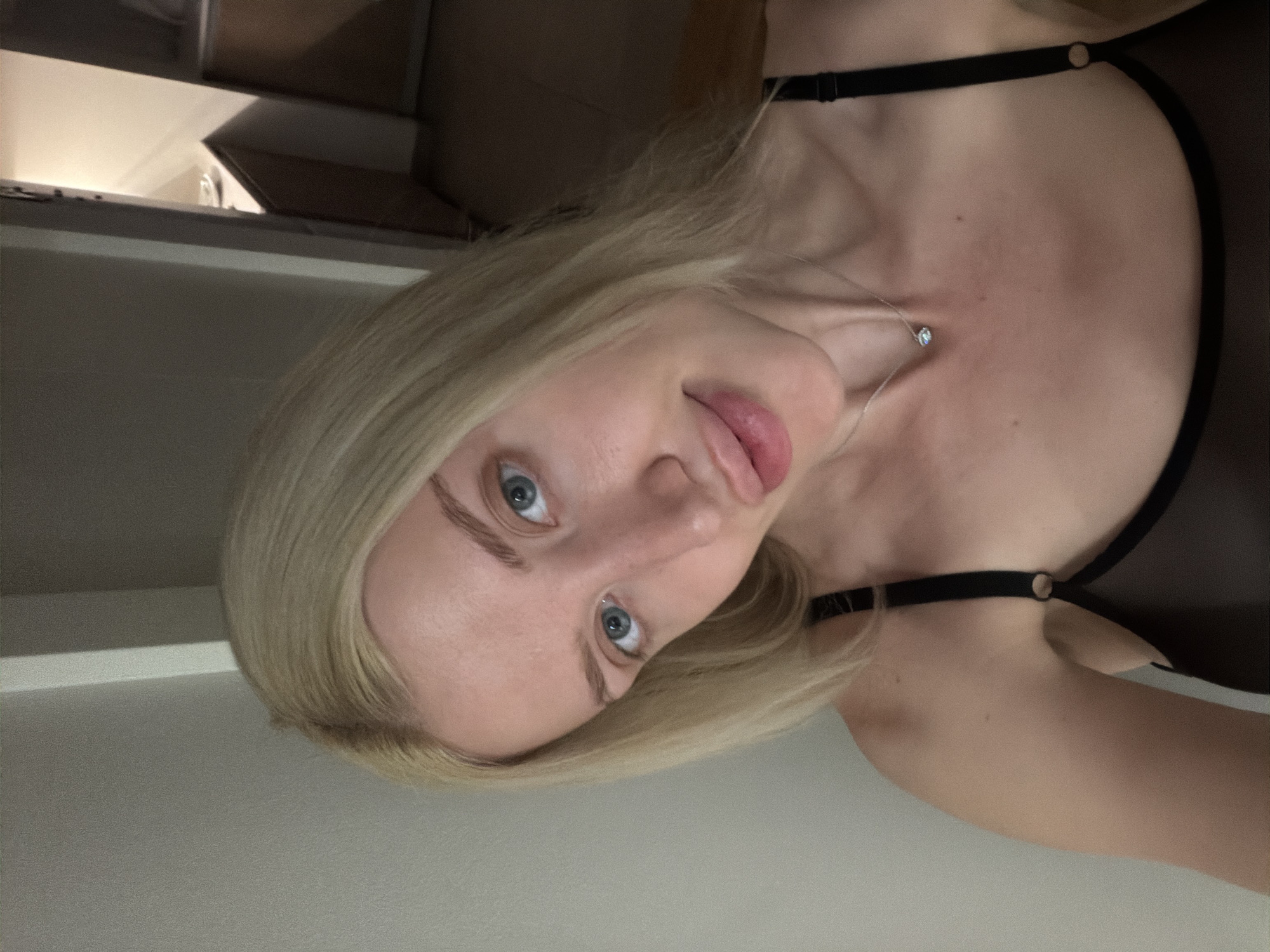 A blonde woman wearing a dark garment with thin straps is facing the camera. She has blue eyes and a silver necklace with a small stone. The background appears to be a neutral-toned room, possibly a domestic interior with a shelf or cabinet visible in the upper left corner. There is no definitive evidence to place this image in Belgrade, Serbia.