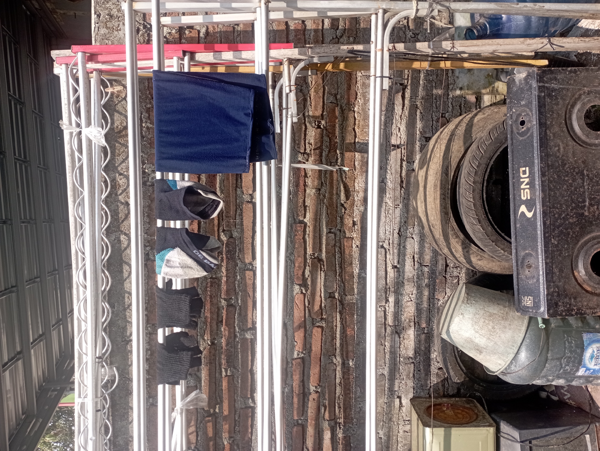The image depicts an outdoor drying rack laden with clothes and laundry items, set against a weathered brick wall. The drying rack is made of white metal tubes, with several items of clothing hanging on it. On the top rack, there are three pairs of dark-colored gloves, followed by a pair of patterned gloves with blue, white, and grey accents. To the right of these gloves, a dark blue towel or piece of fabric is hung.
Below this top rack, the brick wall is visible. The bricks are irregular in shape and color, indicating age and exposure to the elements. Some of the mortar between the bricks is also visible.
The lower portion of the image reveals a collection of objects that appear to be discarded or stored outdoors. There are several tires stacked vertically, one of which is partially obscured by a light grey, dented bucket. The bucket is placed upside down on top of a dark grey plastic container, which has a label with blue lettering. To the left of these items, a light-colored, rectangular object with a round vent is partially visible. In the bottom right corner, a large blue plastic water jug is present. In the foreground, a black, flat object with the "SNG" logo is visible, appearing to be a gas stove or similar appliance.
The scene is brightly lit by sunlight, casting shadows and highlighting the textures of the brick wall and the various objects. The overall impression is one of an outdoor utility area or a backyard storage space. The environment suggests a warm, possibly humid climate, typical of Maos Kidul, Indonesia, where this scene might be found. The presence of household items like laundry, tires, and a water jug points to a residential setting.